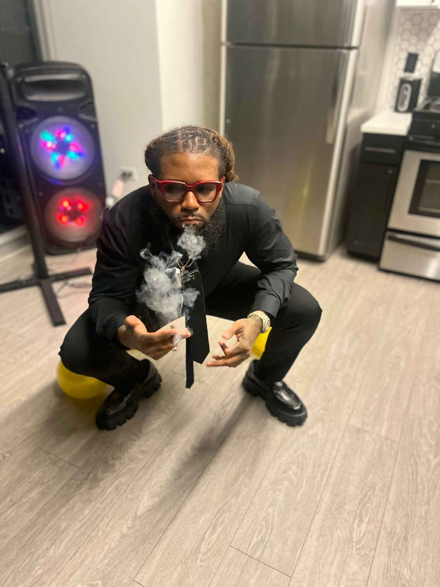 Person crouching in a kitchen, smoking, wearing red sunglasses, black jacket, black pants, and yellow shoes.
