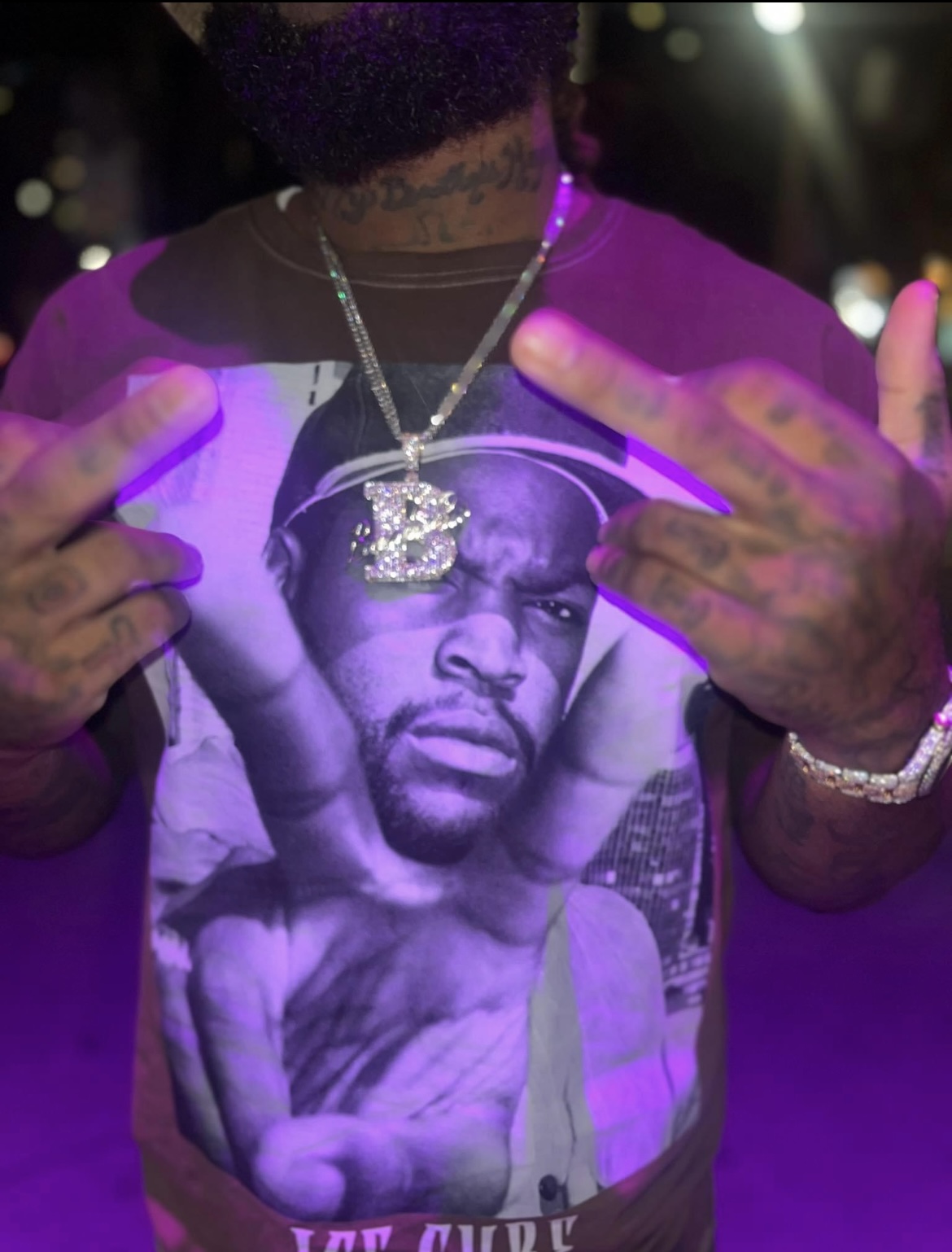 Person wearing a graphic T-shirt with a black-and-white image of a man, holding up two fingers near their face, with jewelry and tattoos.