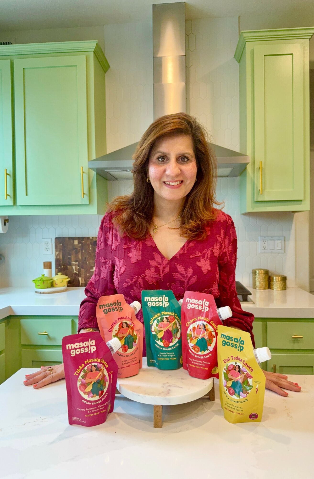 Woman standing in a kitchen with green cabinets, smiling, with colorful snack packages on a tray in front.