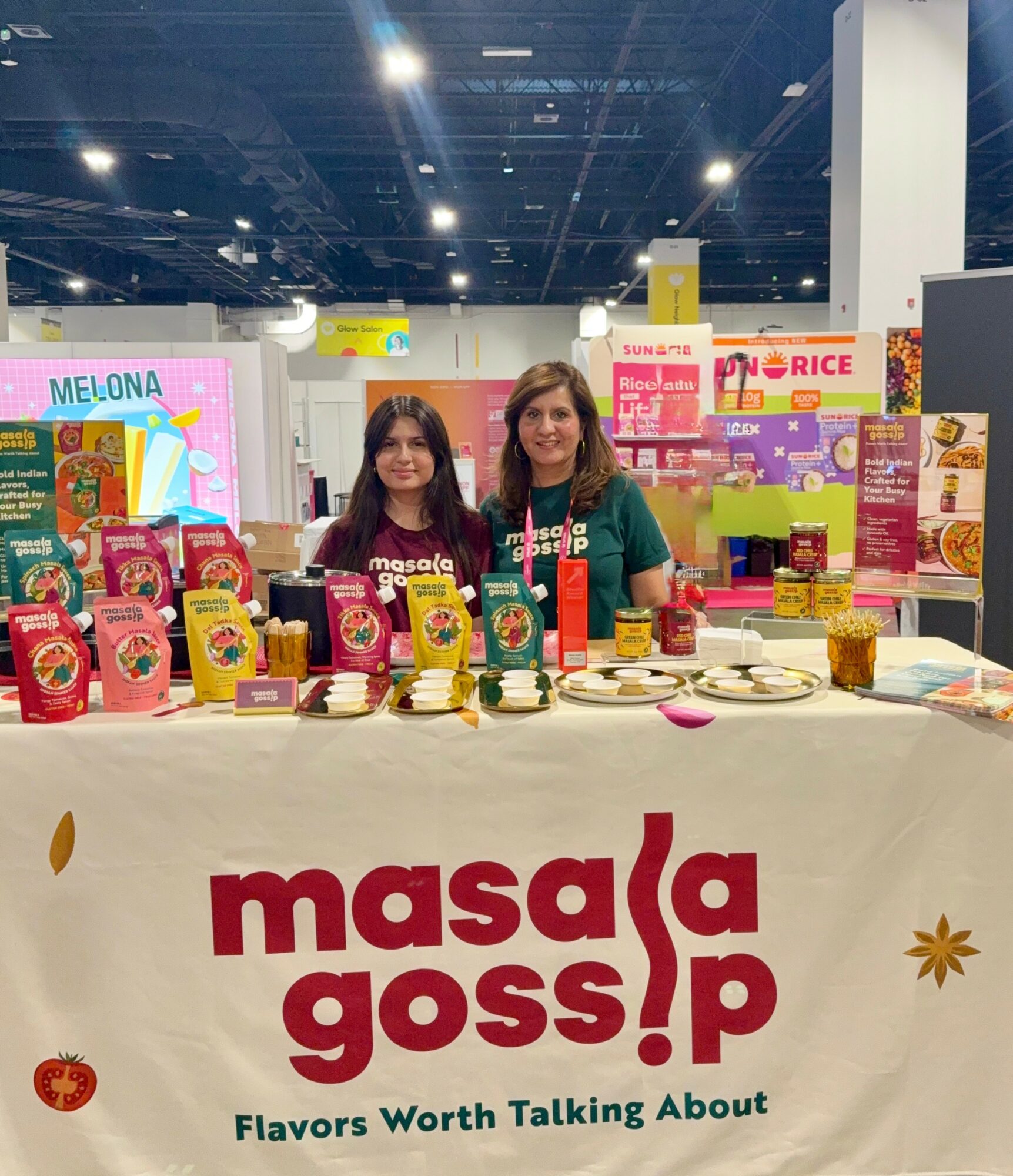 Two women stand behind a table displaying various packaged food products at a trade show booth with colorful signage and branding.