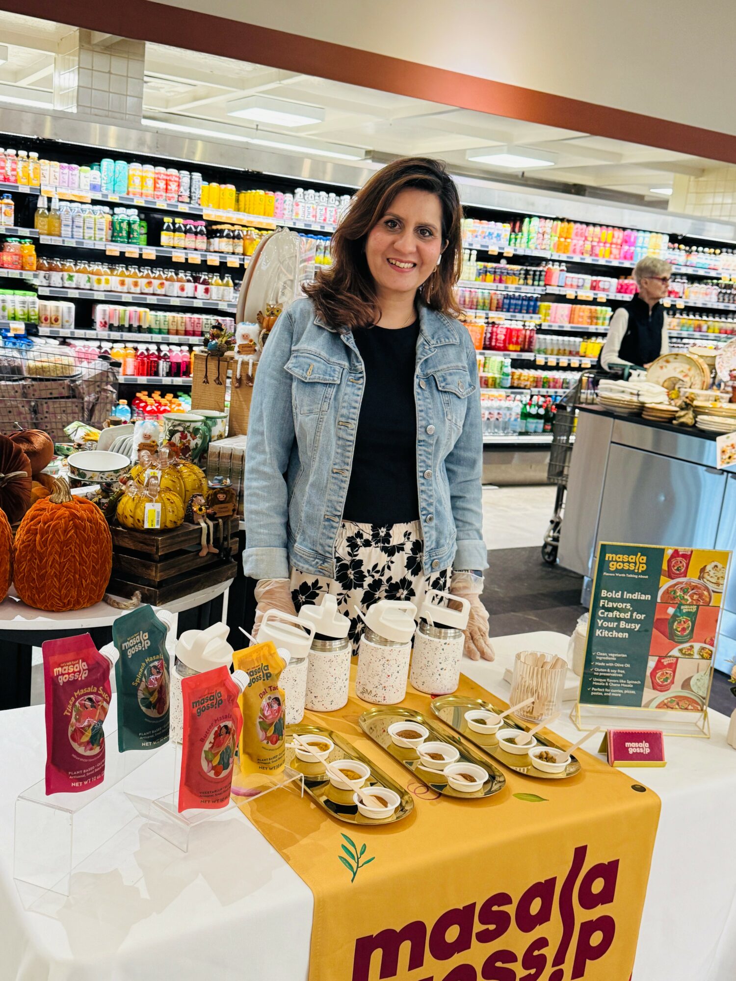 Woman standing behind a table with jars and snacks in a store aisle, smiling, with shelves of products behind her.