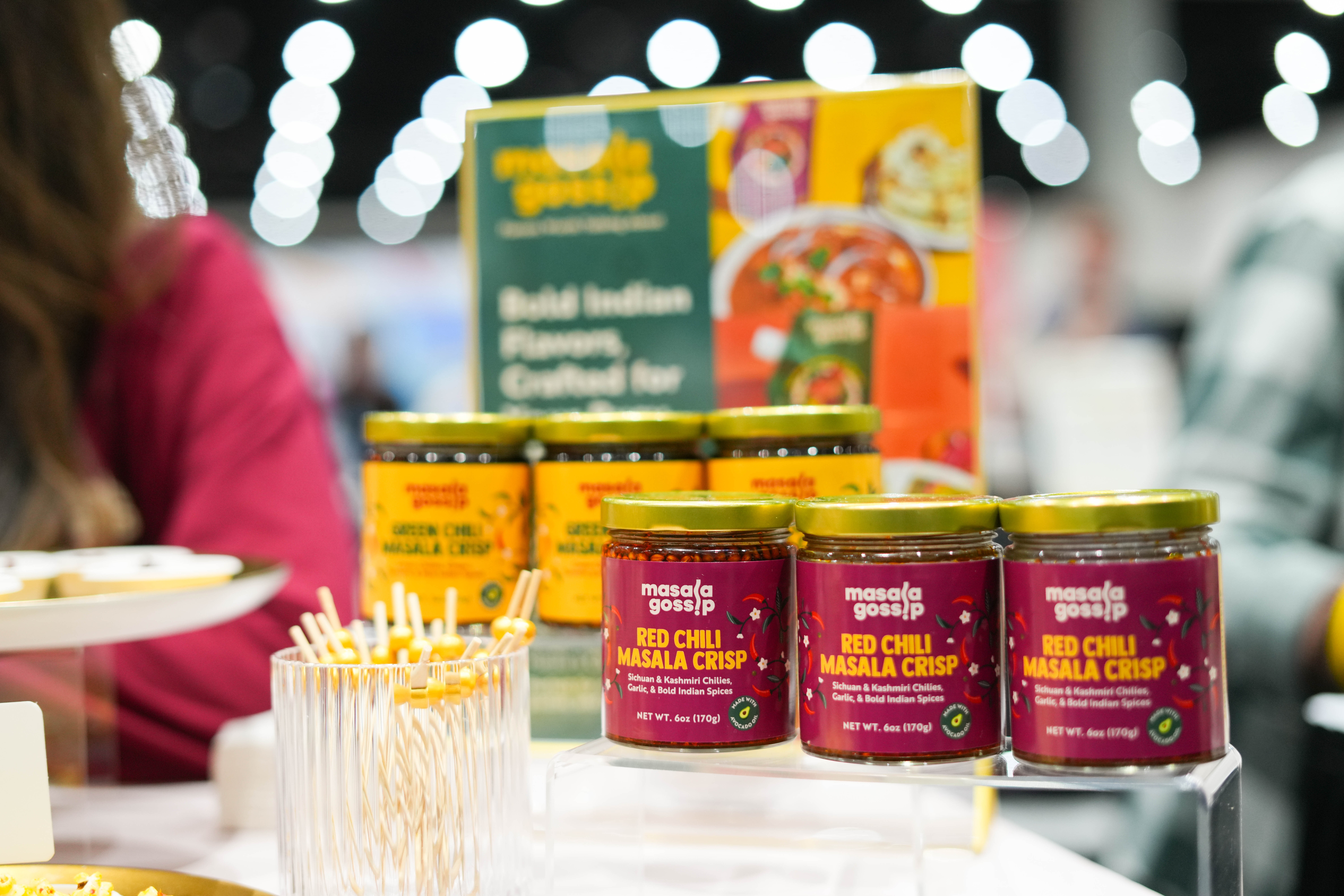 Display of jars with red chili label and a colorful menu board in the background at a market or store, blurred background.