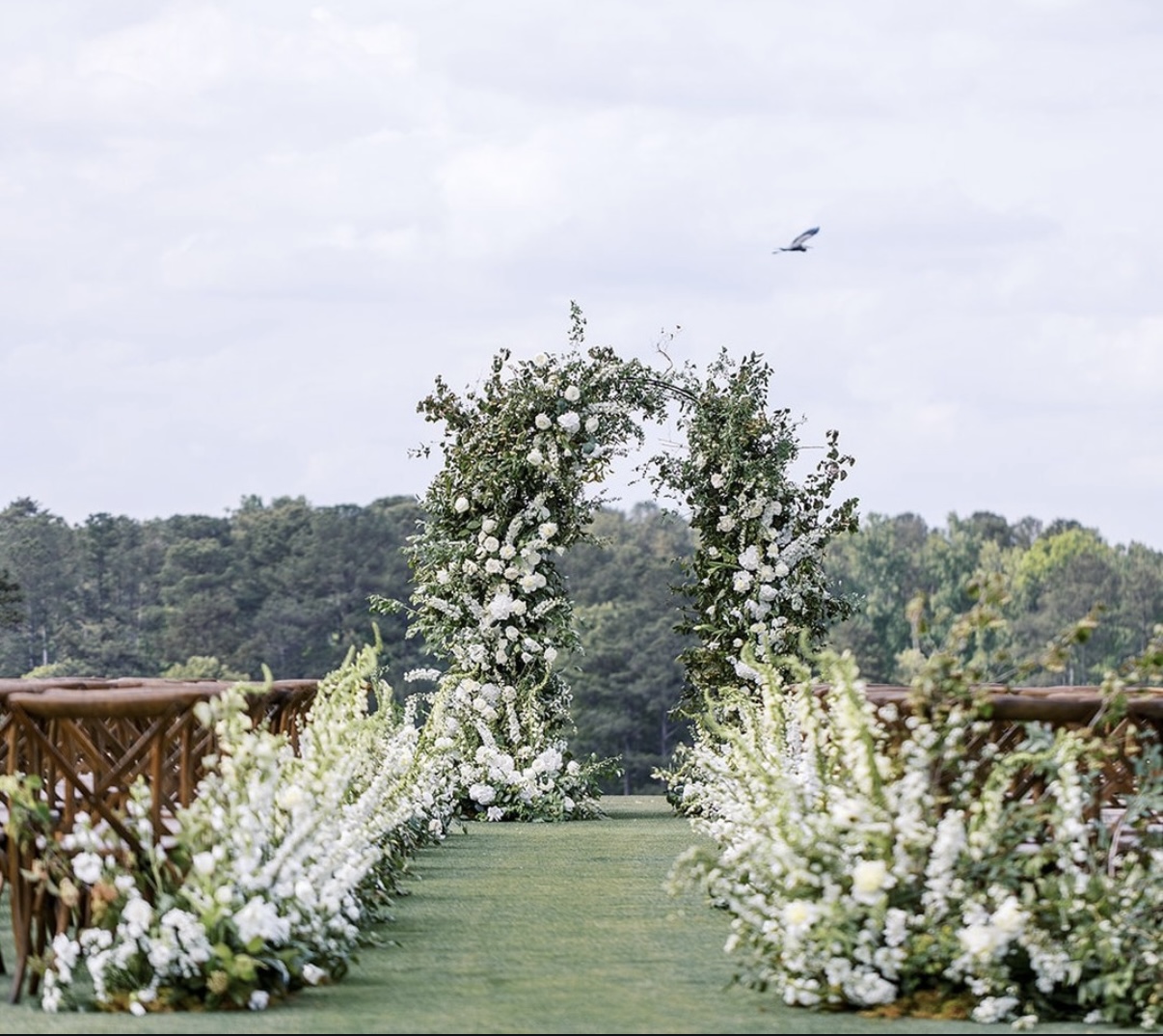 Flower arch with white flowers and greenery over a grassy aisle, outdoor setting with trees and cloudy sky