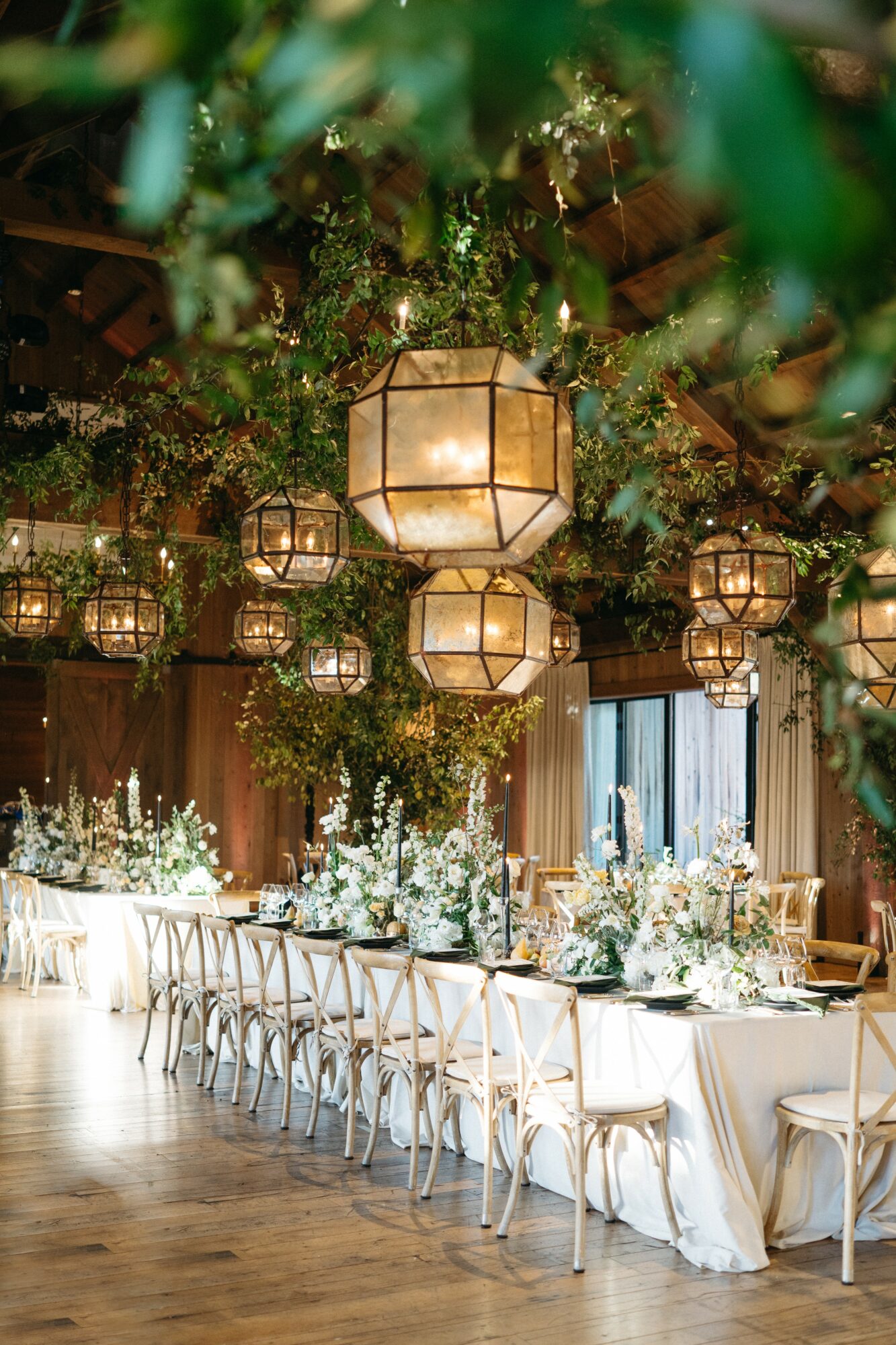 Elegant banquet table with white chairs, floral centerpieces, and hanging lanterns in a decorated event space.