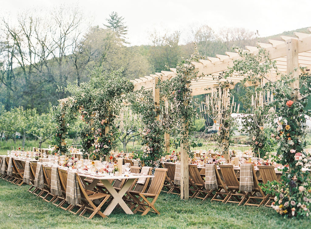 Long outdoor table set with chairs, decorated with flowers and greenery, in a garden setting with trees and a building in background.