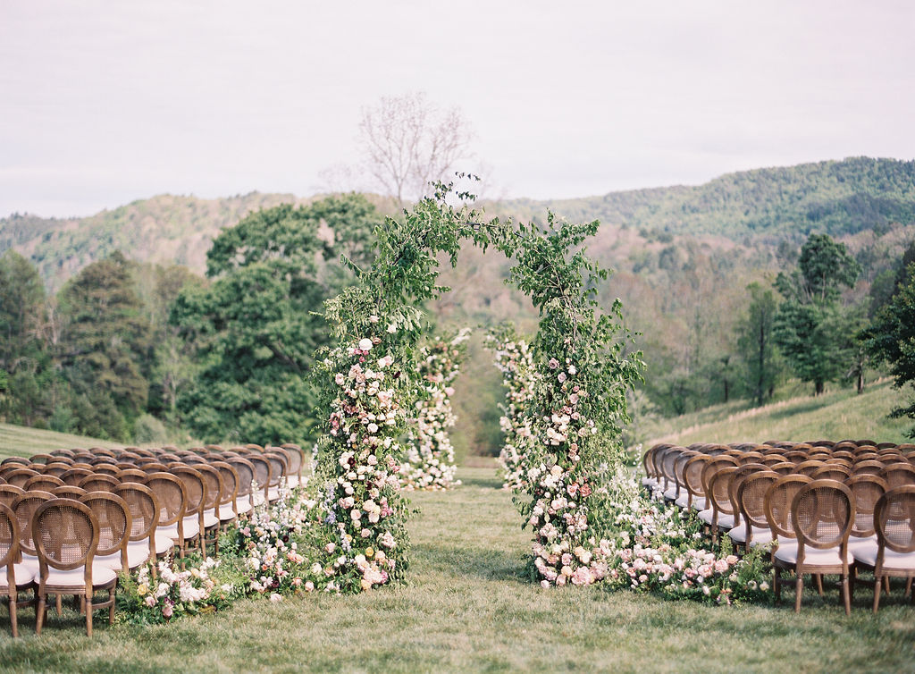 Outdoor wedding setup with floral arch and rows of chairs on grass, trees, and hills in background.