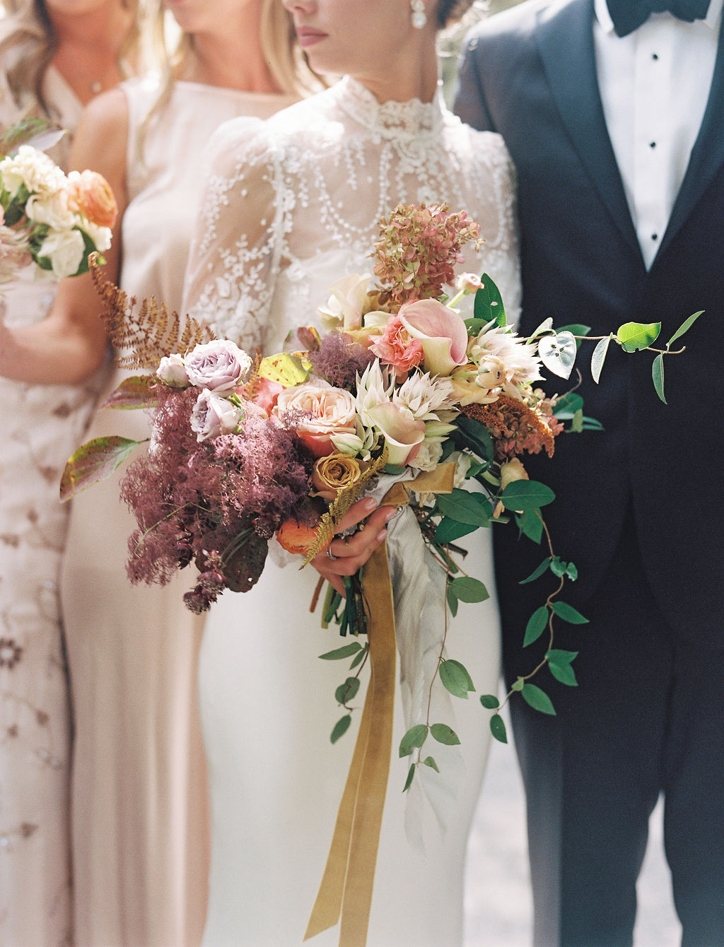 Bride holding a large bouquet of pink, cream, and purple flowers with greenery, surrounded by people in formal attire.