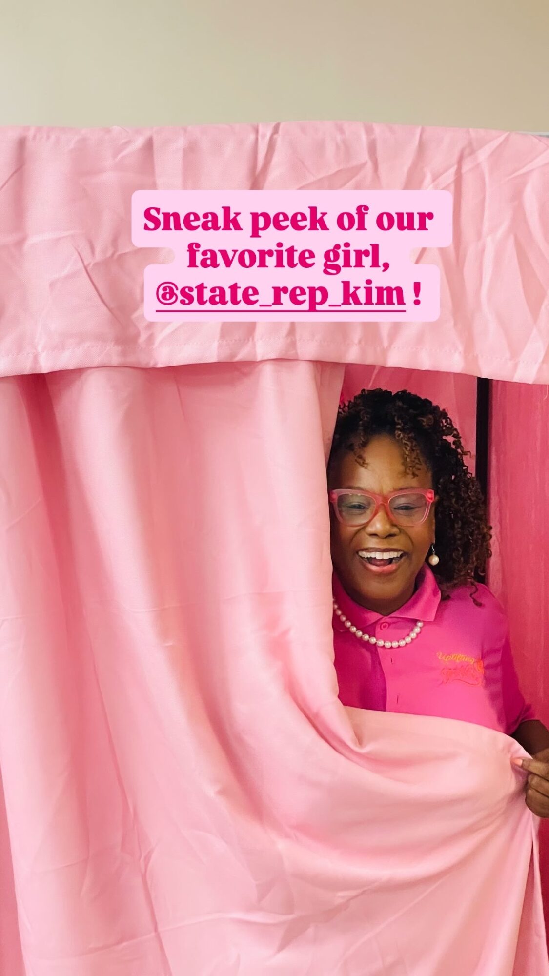 Person with curly hair and glasses peeking through pink curtains, smiling, wearing a pink top, with a pink banner above.