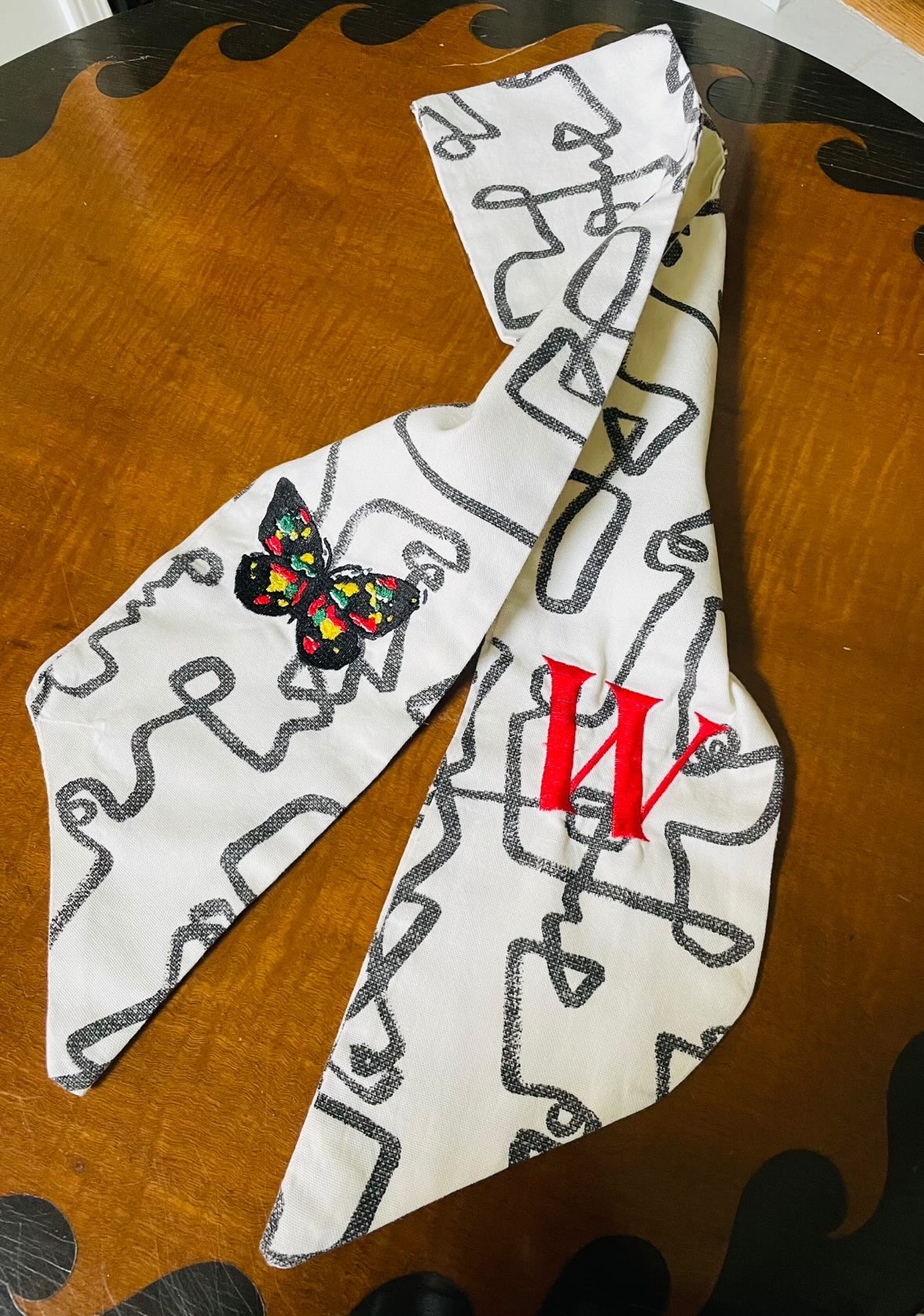 A fabric with black scribbles, a colorful butterfly patch, and red initials, draped over a wooden surface.