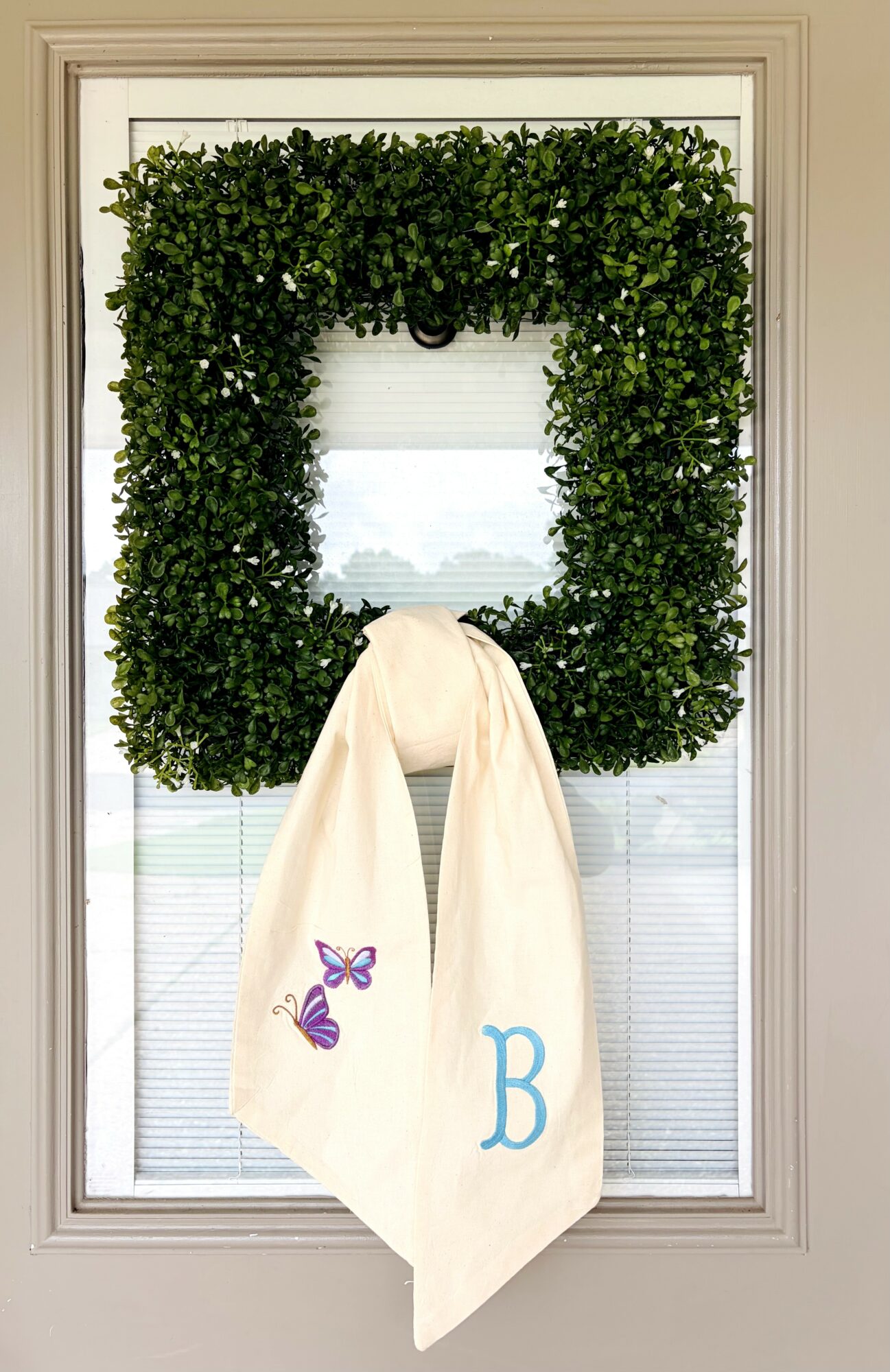 Decorative window with a green square wreath and a white towel with a purple butterfly and blue letter B hanging on it.