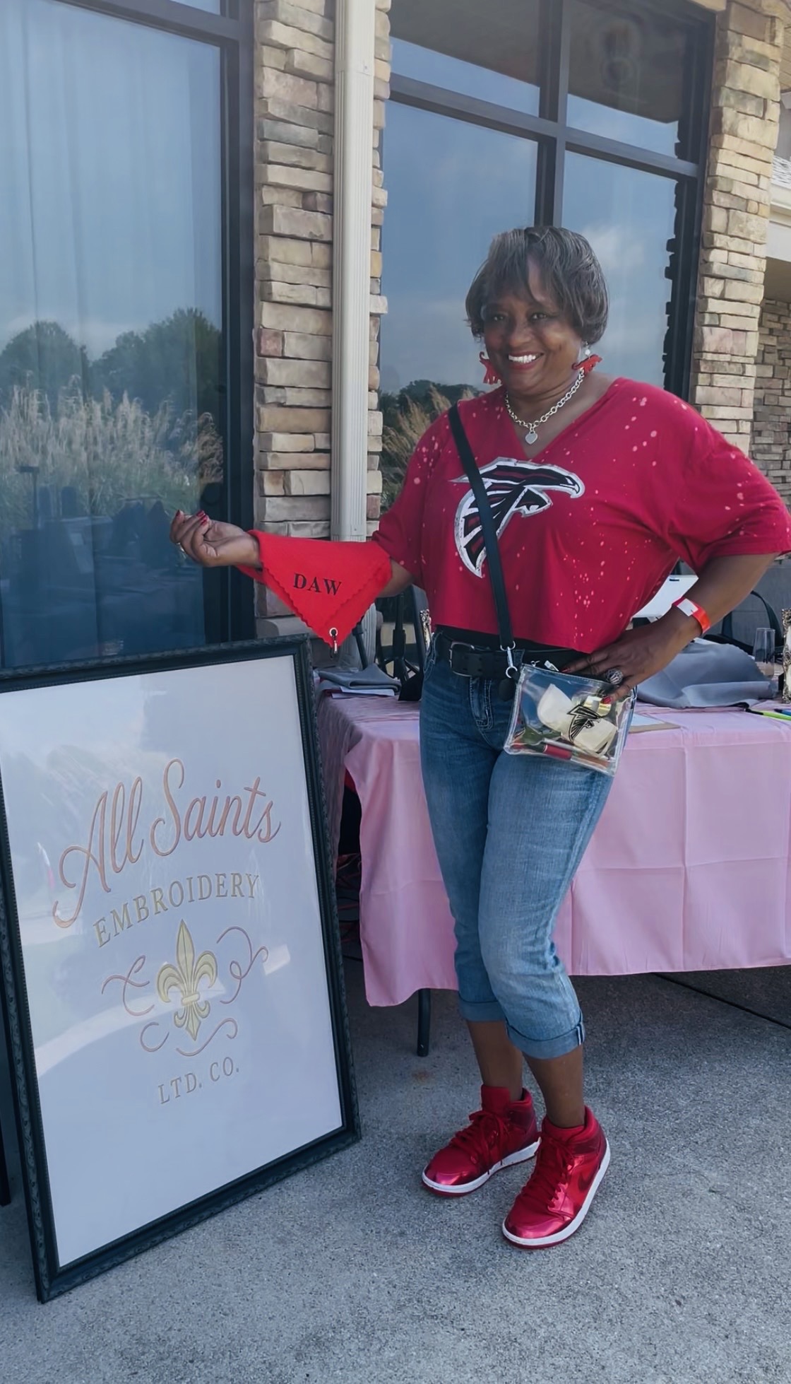 Woman in red top and jeans standing outside next to a sign, smiling, with a building and window behind her.