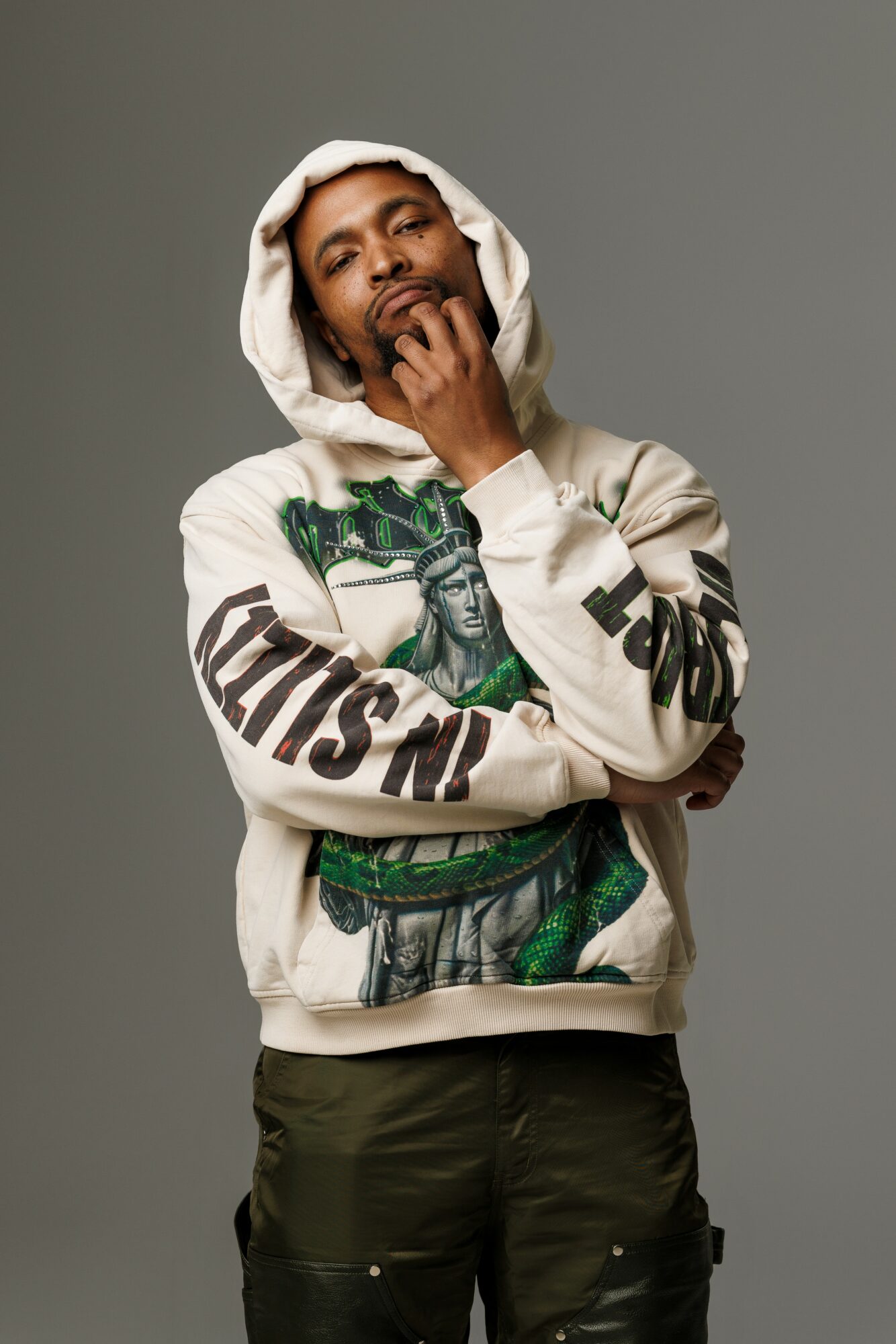 Young man with a hoodie, hand near mouth, wearing a graphic sweatshirt with a statue and text, standing against a gray background.