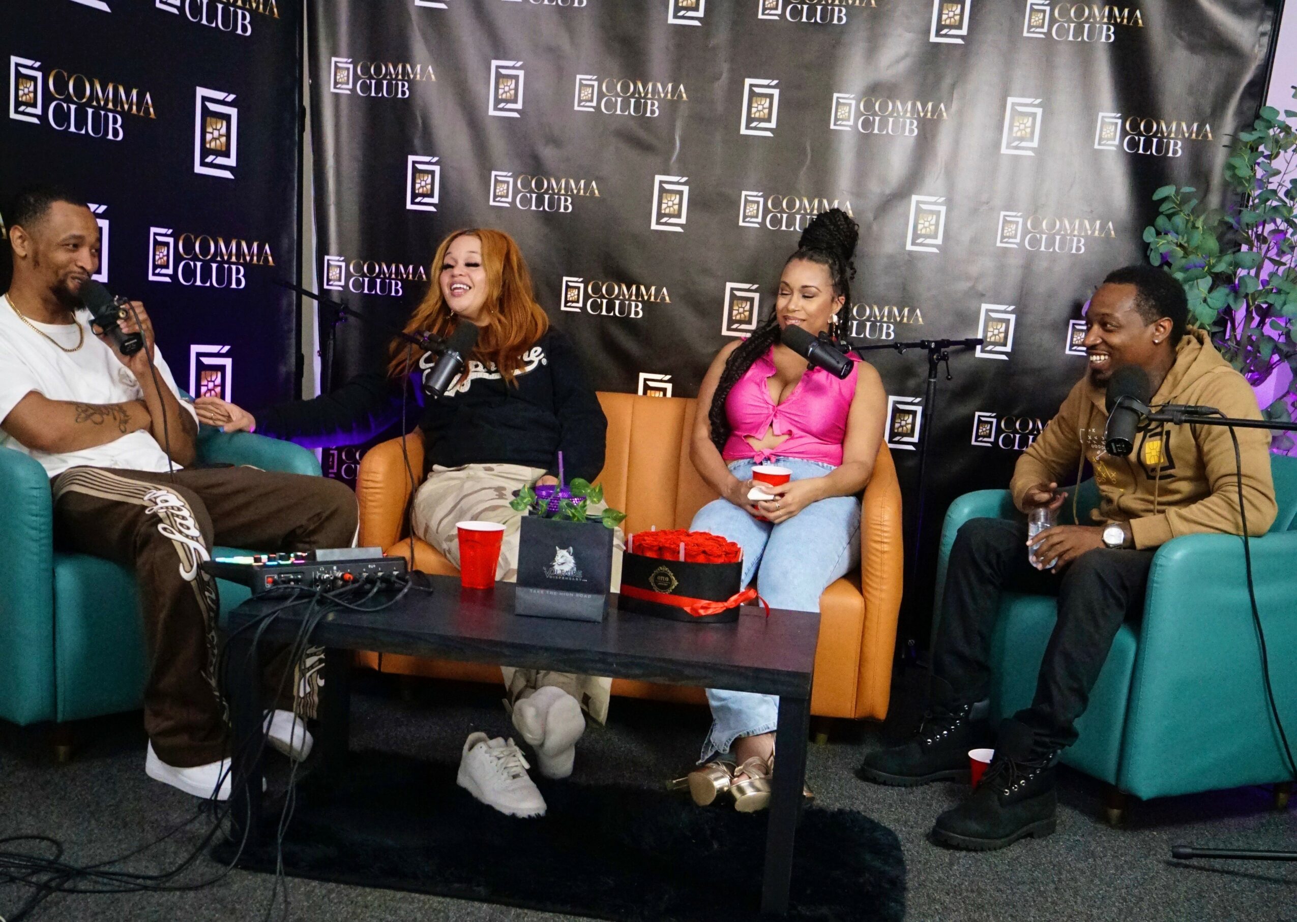Four people sitting on colorful chairs in a talk show setting with a black backdrop and purple flowers, engaging in conversation.