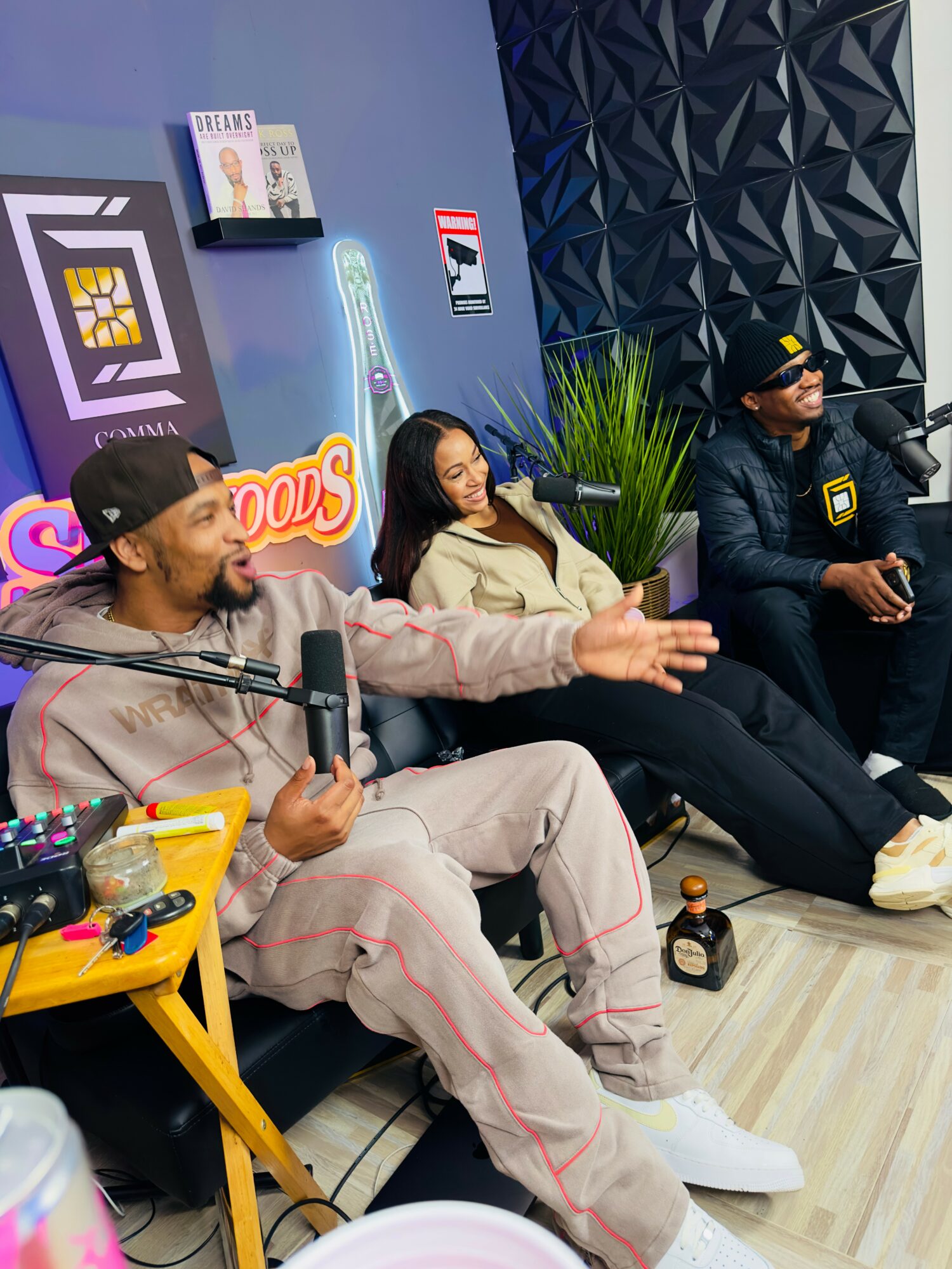 Three people sit on a couch in a room with decorated walls, one speaking into a microphone, others listening, plants nearby.