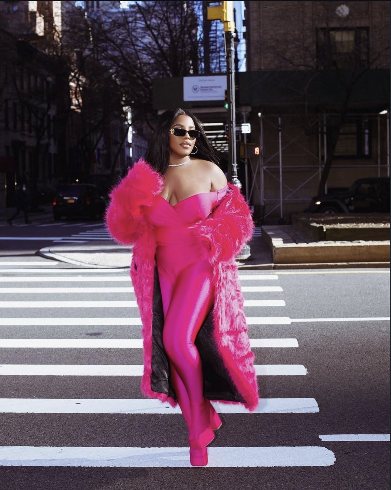 Woman in pink outfit and sunglasses standing on crosswalk in city street.