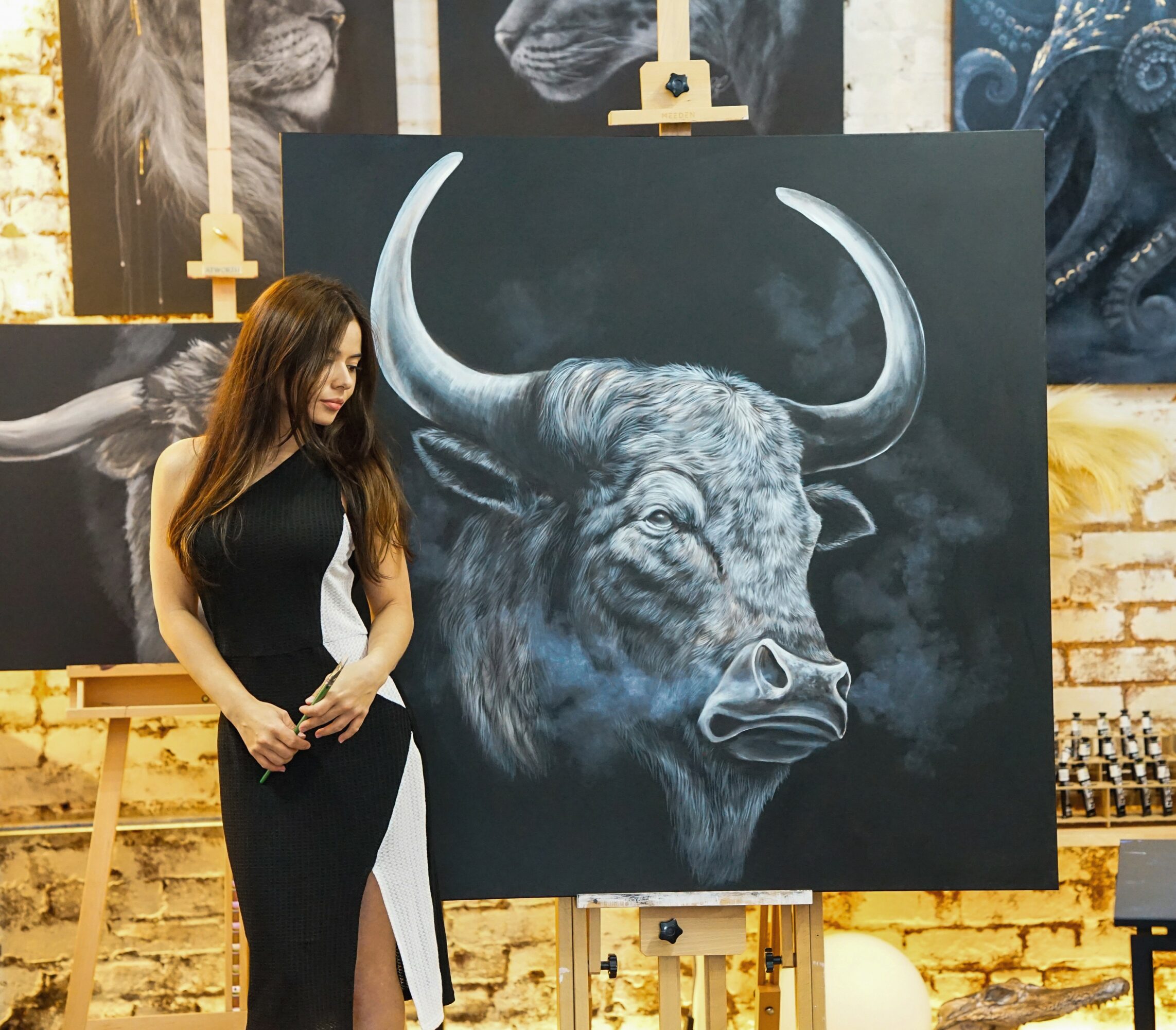 Woman with long hair in black dress stands next to a large painting of a buffalo's head on an easel.