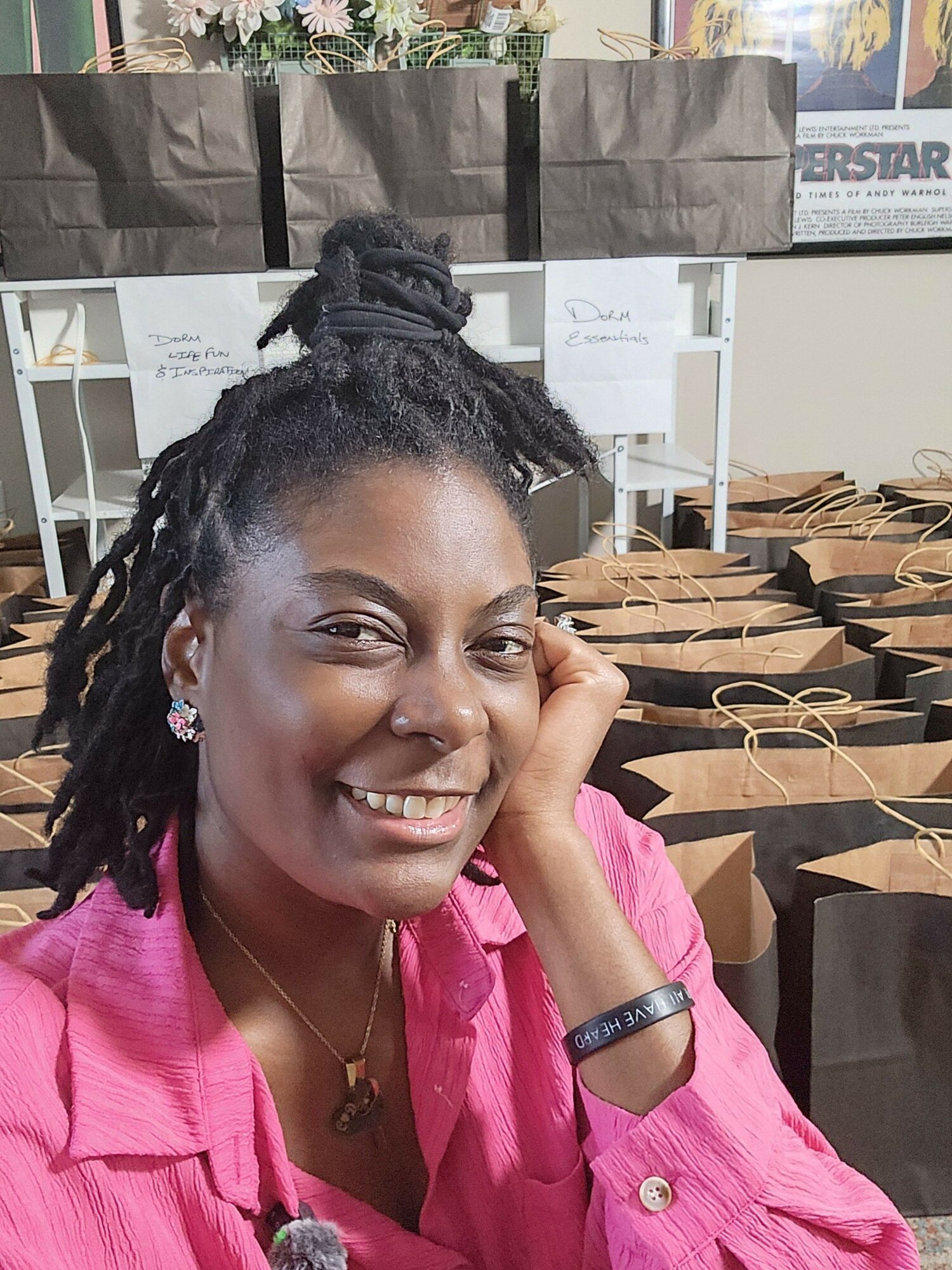 Smiling woman with dreadlocks resting her head on her hand, wearing a pink shirt, outdoors with tables and umbrellas behind her.