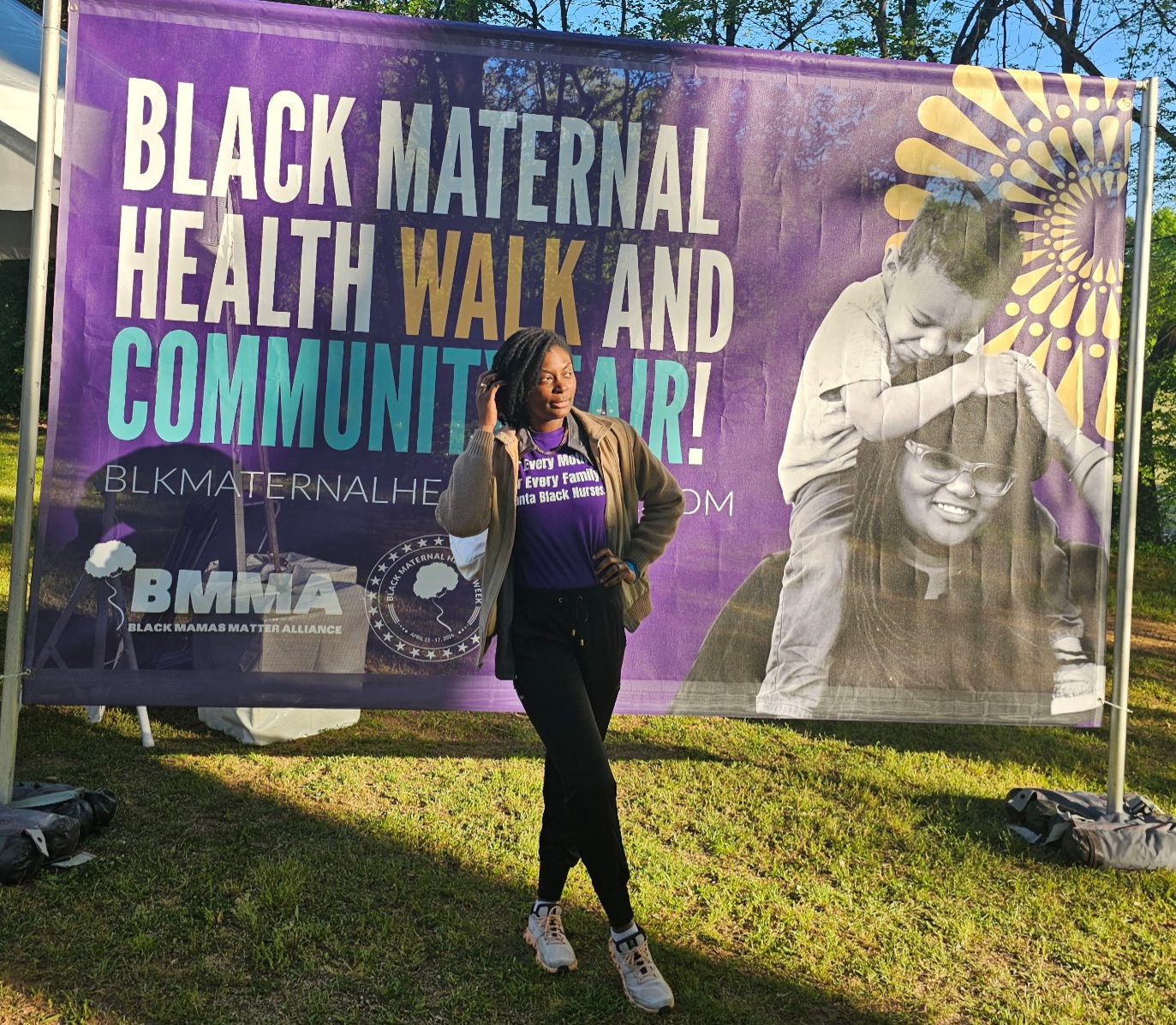 Woman standing outdoors in front of a large purple banner promoting Black maternal health and community, with images of women and children.