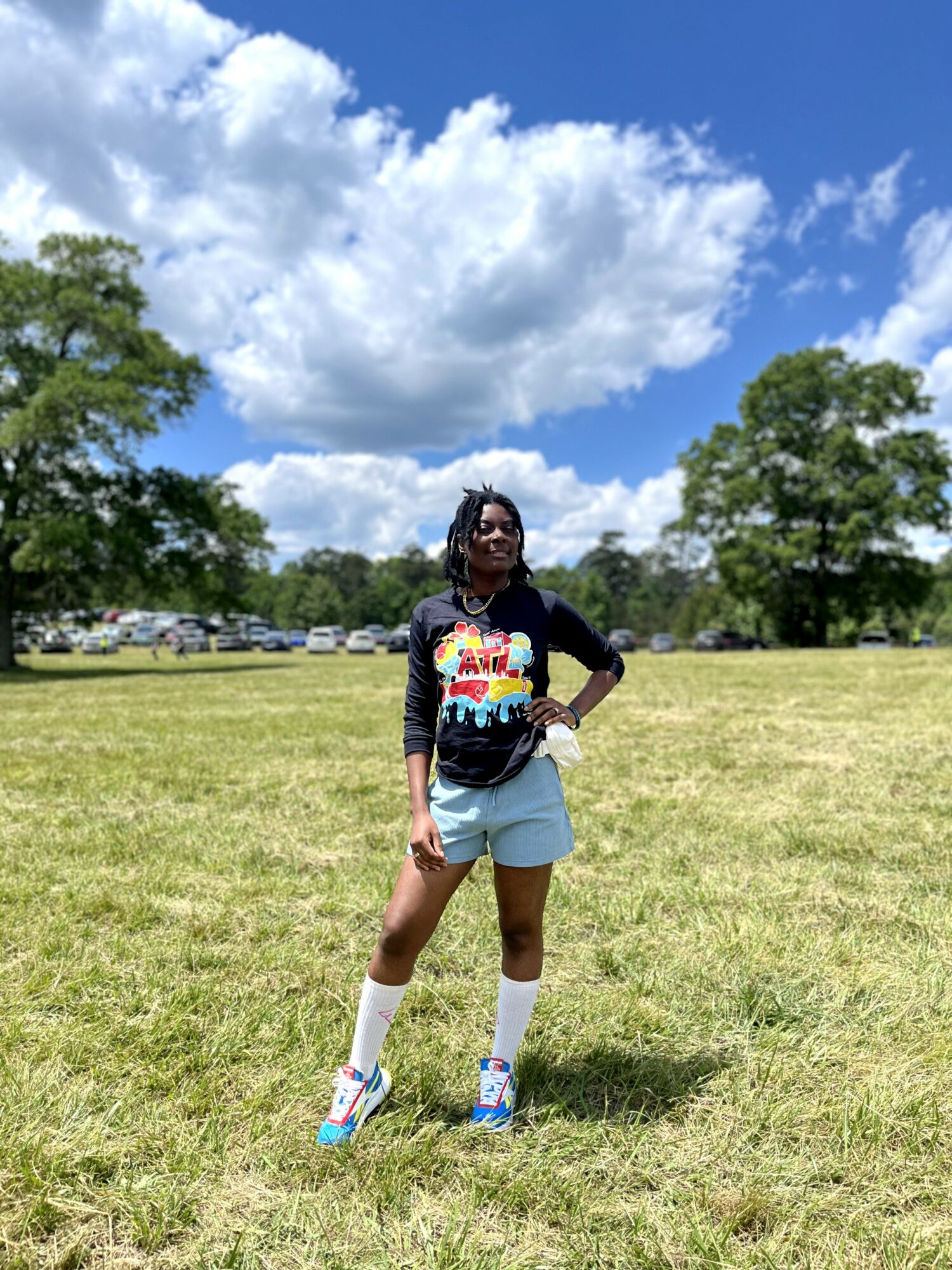 Person standing on grassy field with trees and blue sky with clouds in background, wearing a colorful shirt, shorts, and sneakers.
