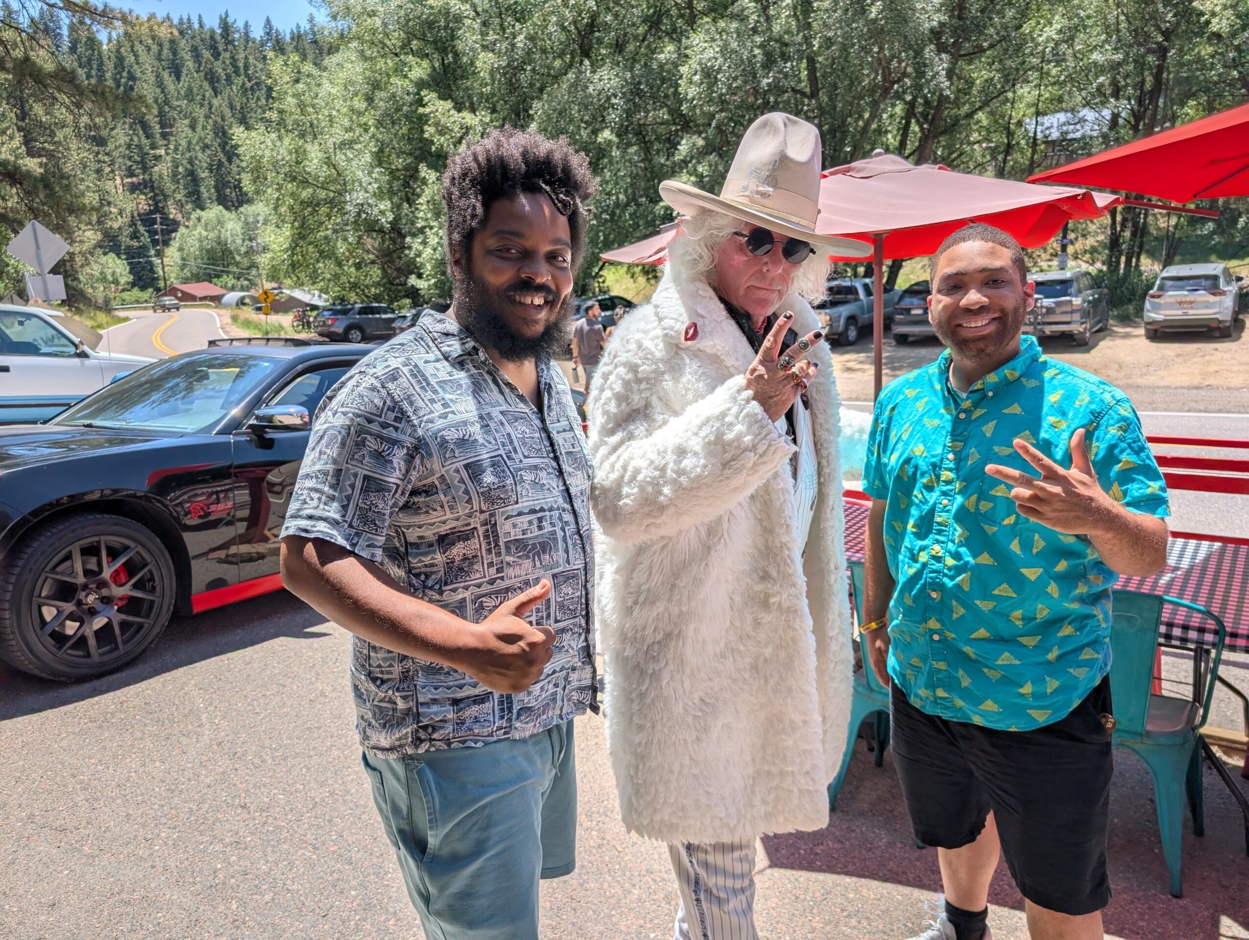 Three people outdoors, one in a white coat and hat, two smiling men, trees and cars in background.