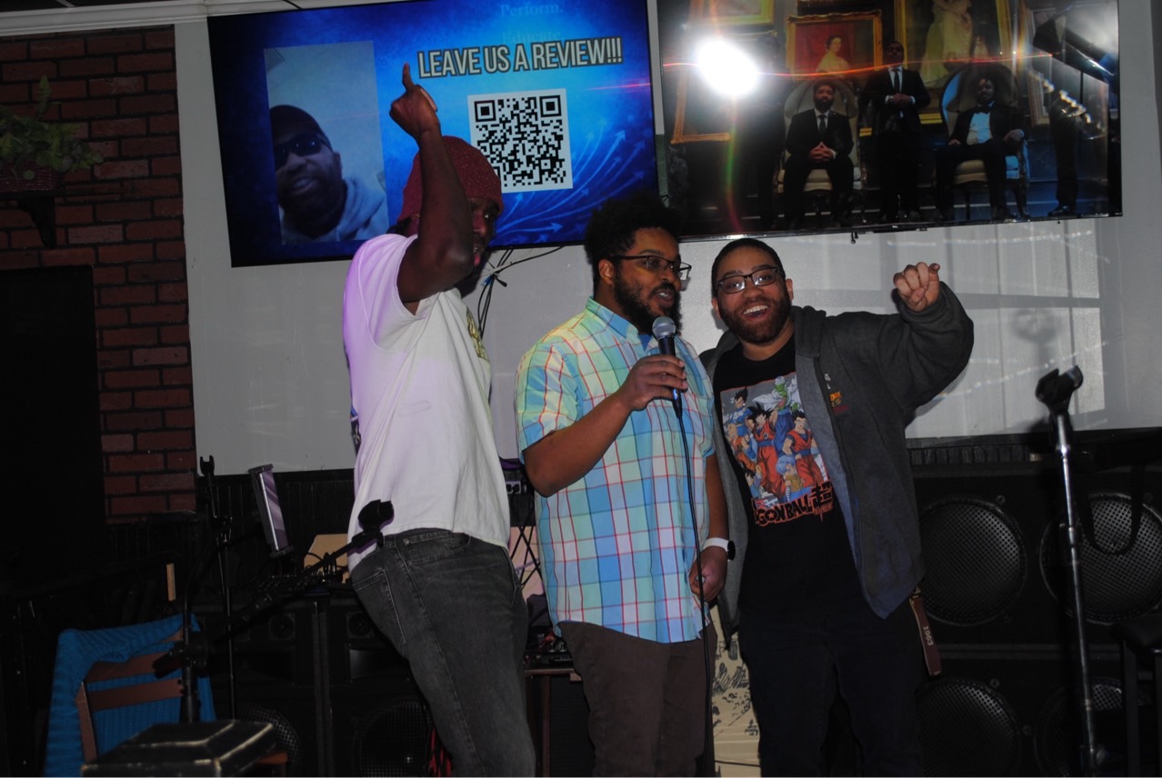 Three men stand together, one holding a microphone, in a dimly lit indoor setting with screens behind them.