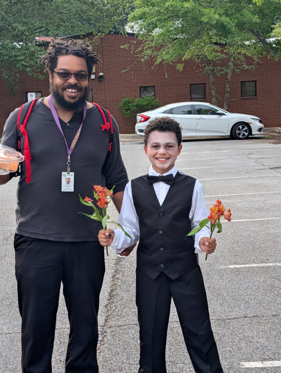 A man and a boy stand outdoors in a parking lot, holding flowers and smiling. The man wears glasses and a backpack, the boy is dressed in a tuxedo with a bow tie.