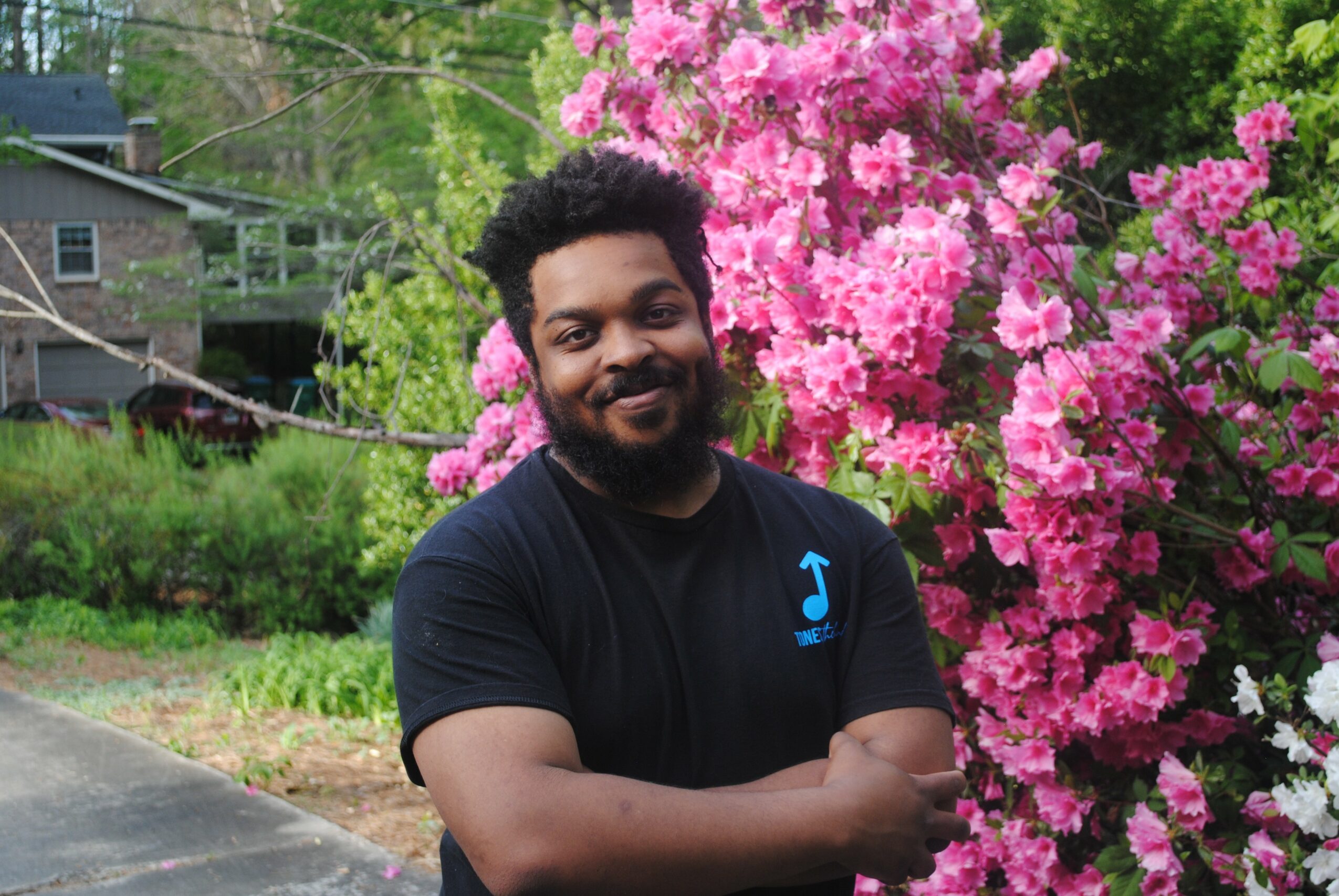 Man with arms crossed standing next to pink flowering bush outdoors, smiling, in a residential area.