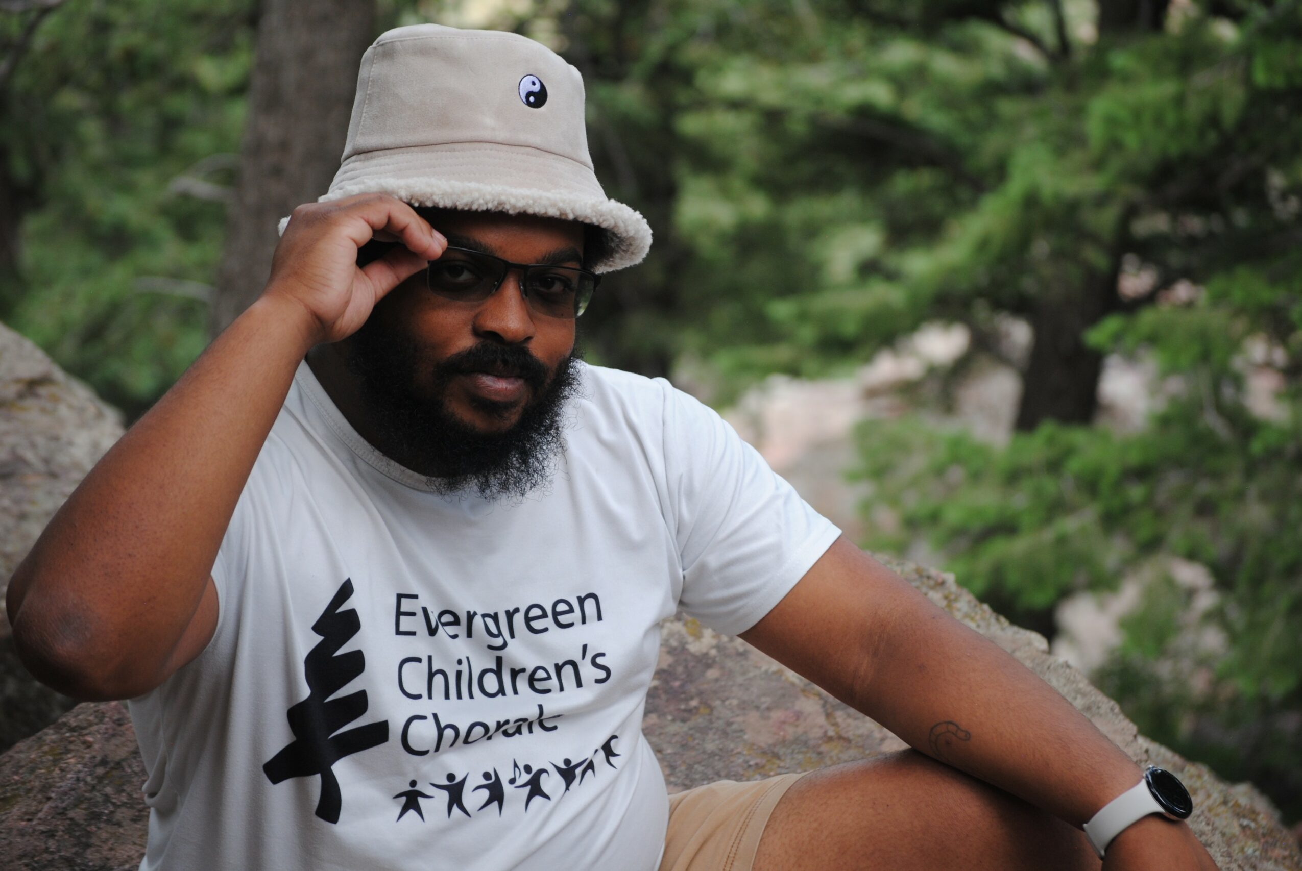Person wearing a bucket hat and sunglasses, sitting outdoors with trees in the background.