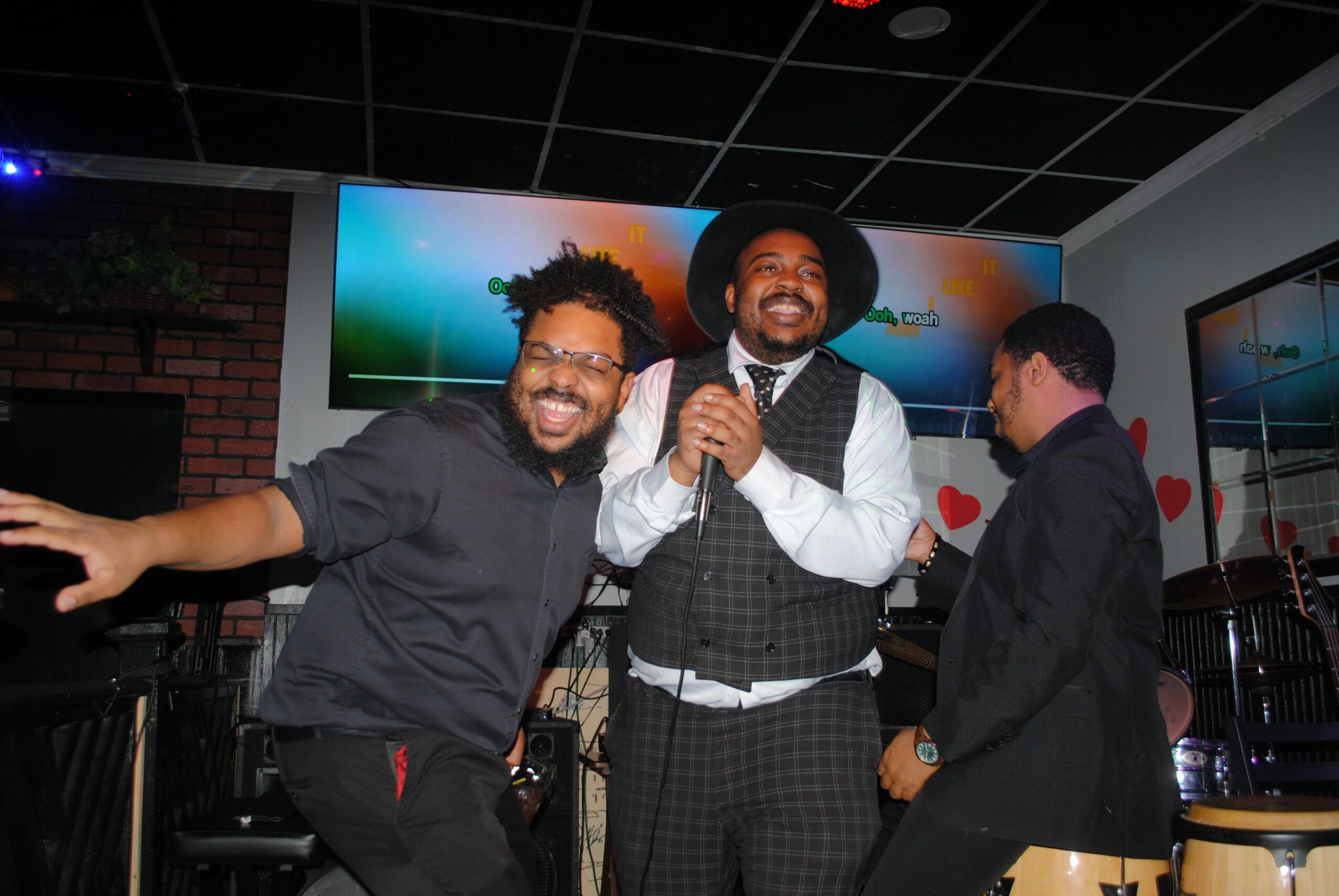 Three men smiling and singing together in a room with screens and hearts on the wall.