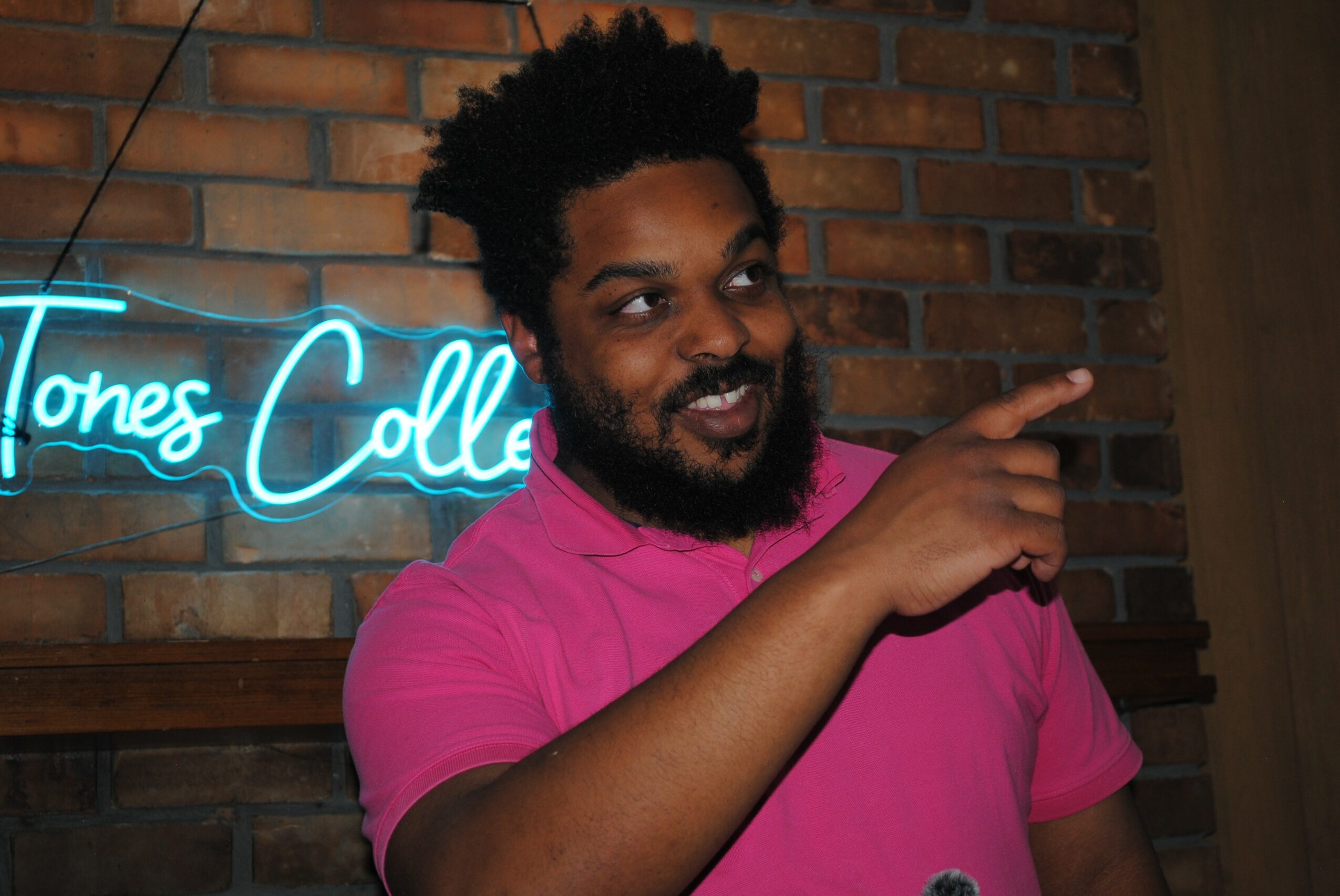 Man with curly hair and beard wearing a pink shirt, pointing to the right, standing in front of a brick wall with a neon sign.