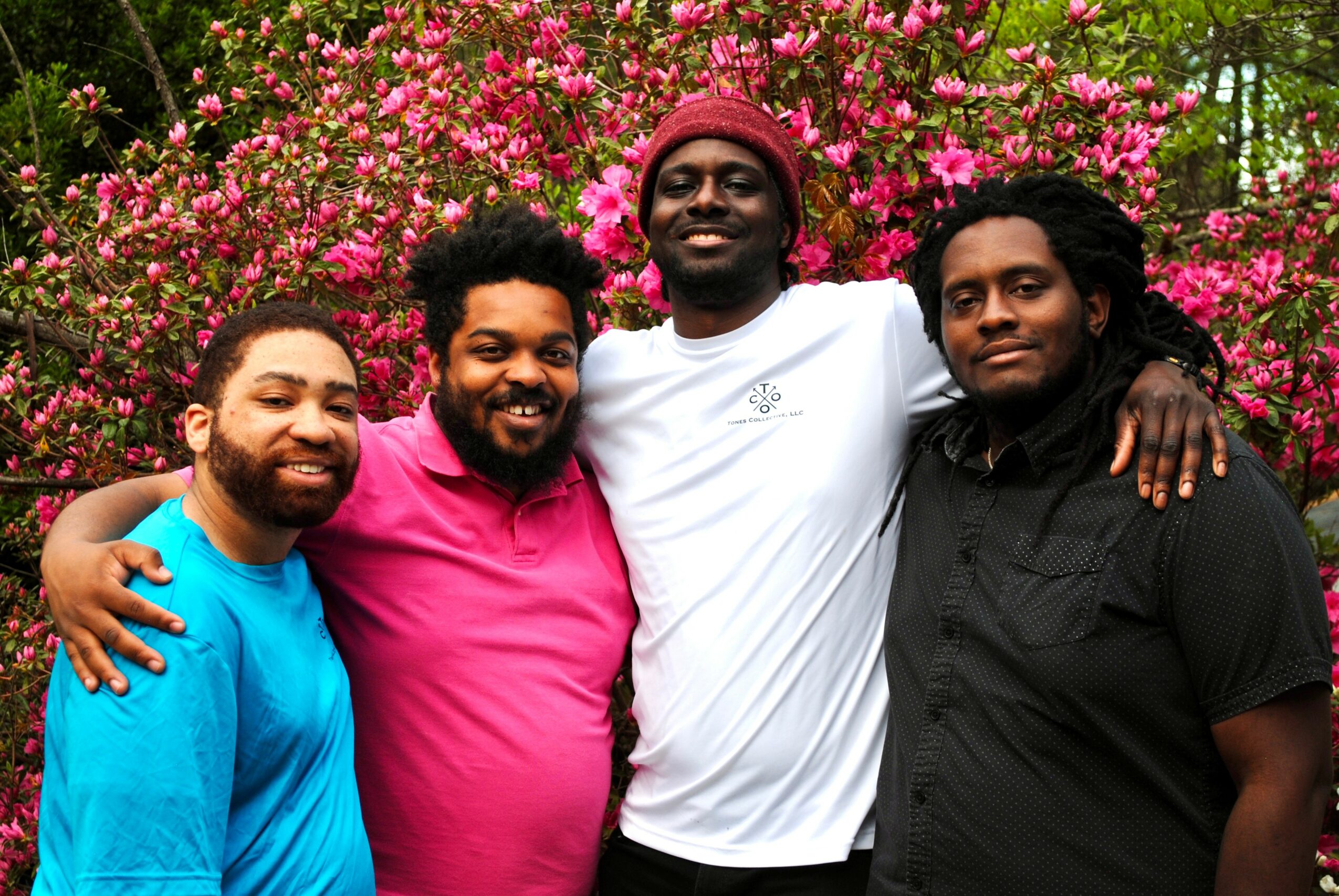 Four people standing outdoors in front of pink flowering bushes, smiling, with arms around each other.