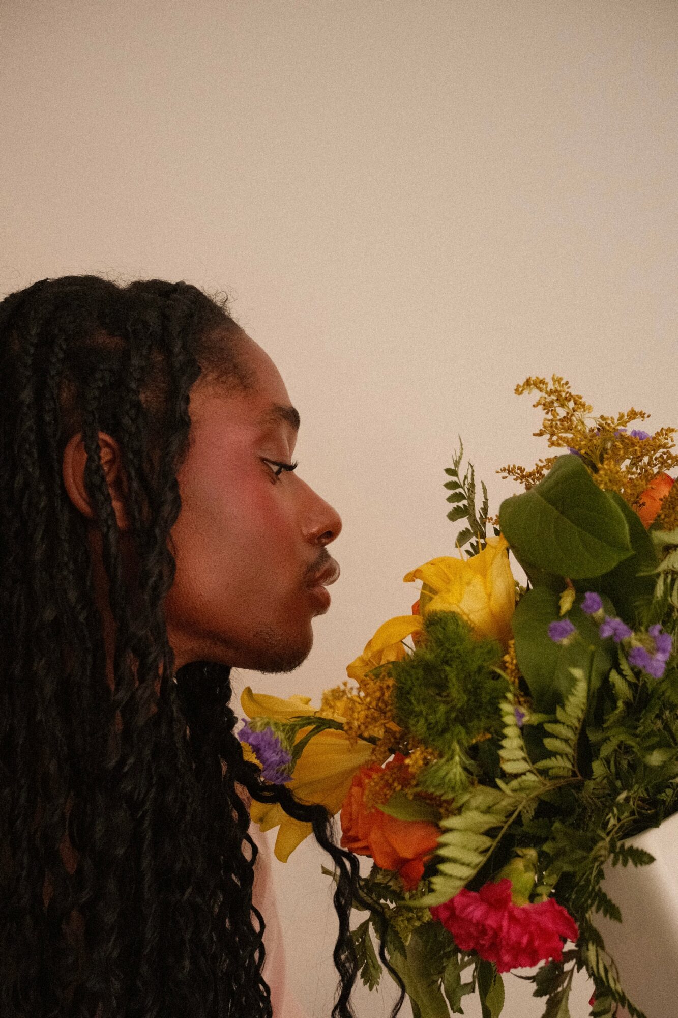 Profile of a woman with long, dark, curly hair looking at a colorful bouquet of flowers against a plain background.