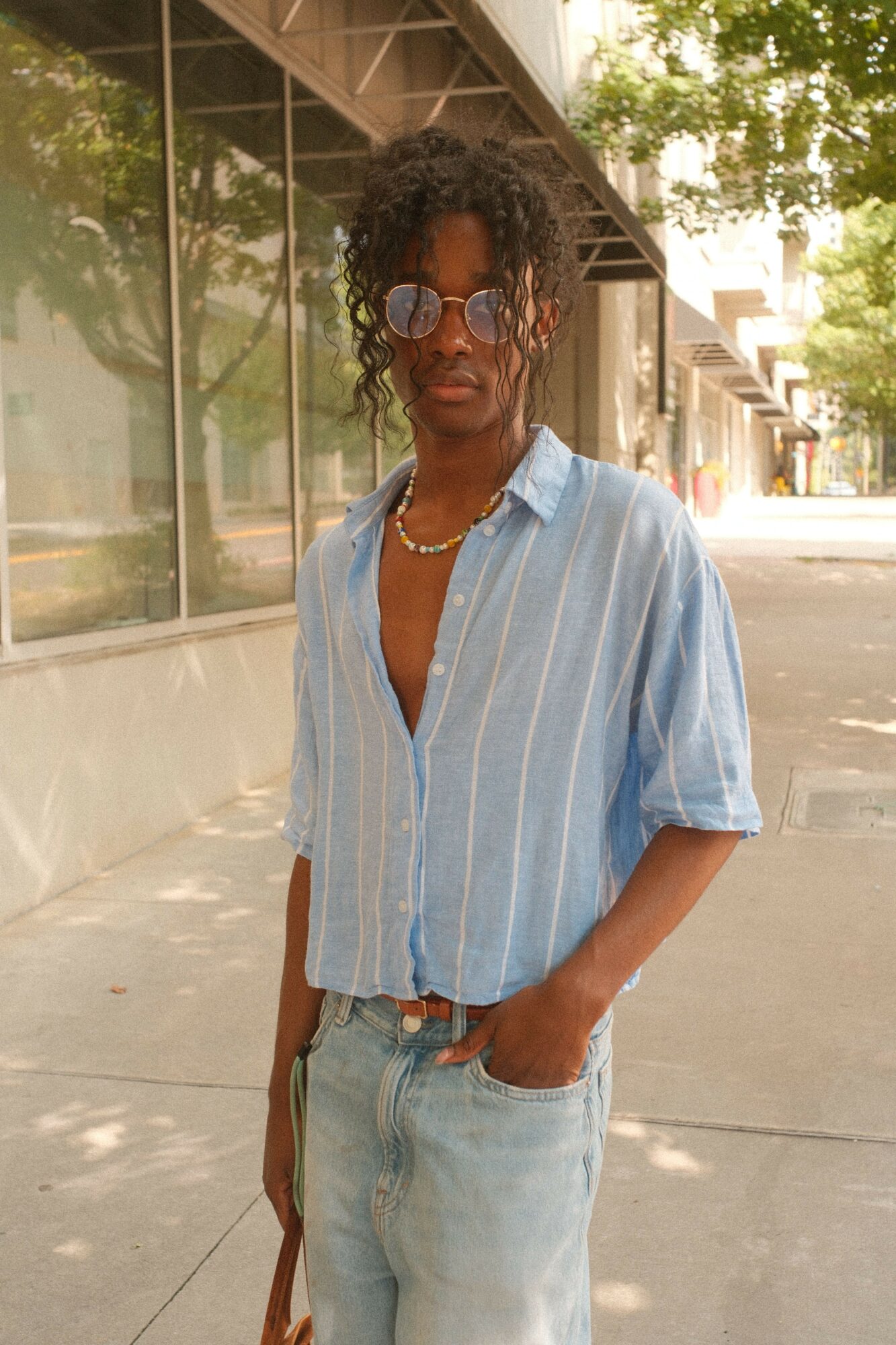 Woman with curly hair and glasses standing outdoors near glass building, wearing a light blue striped shirt and jeans.