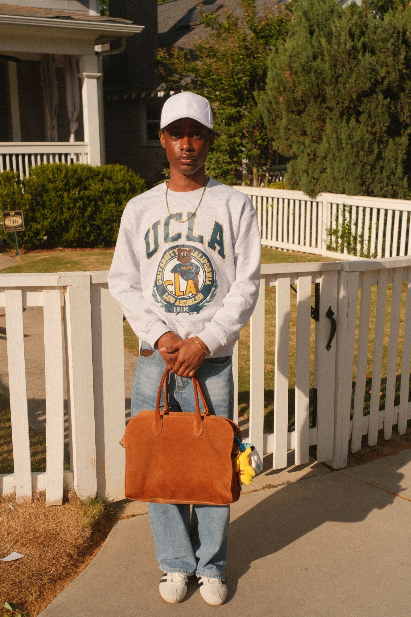 Person standing outdoors holding a large brown bag, wearing a white UCLA sweatshirt and white cap, with a white fence and greenery in background.