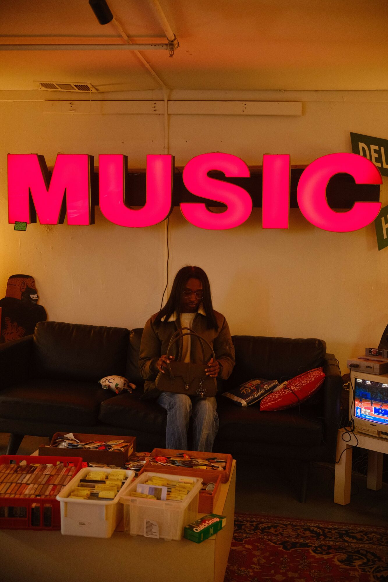Person sitting on a black couch in front of a large pink 'MUSIC' sign, with a table nearby, in a store or venue.