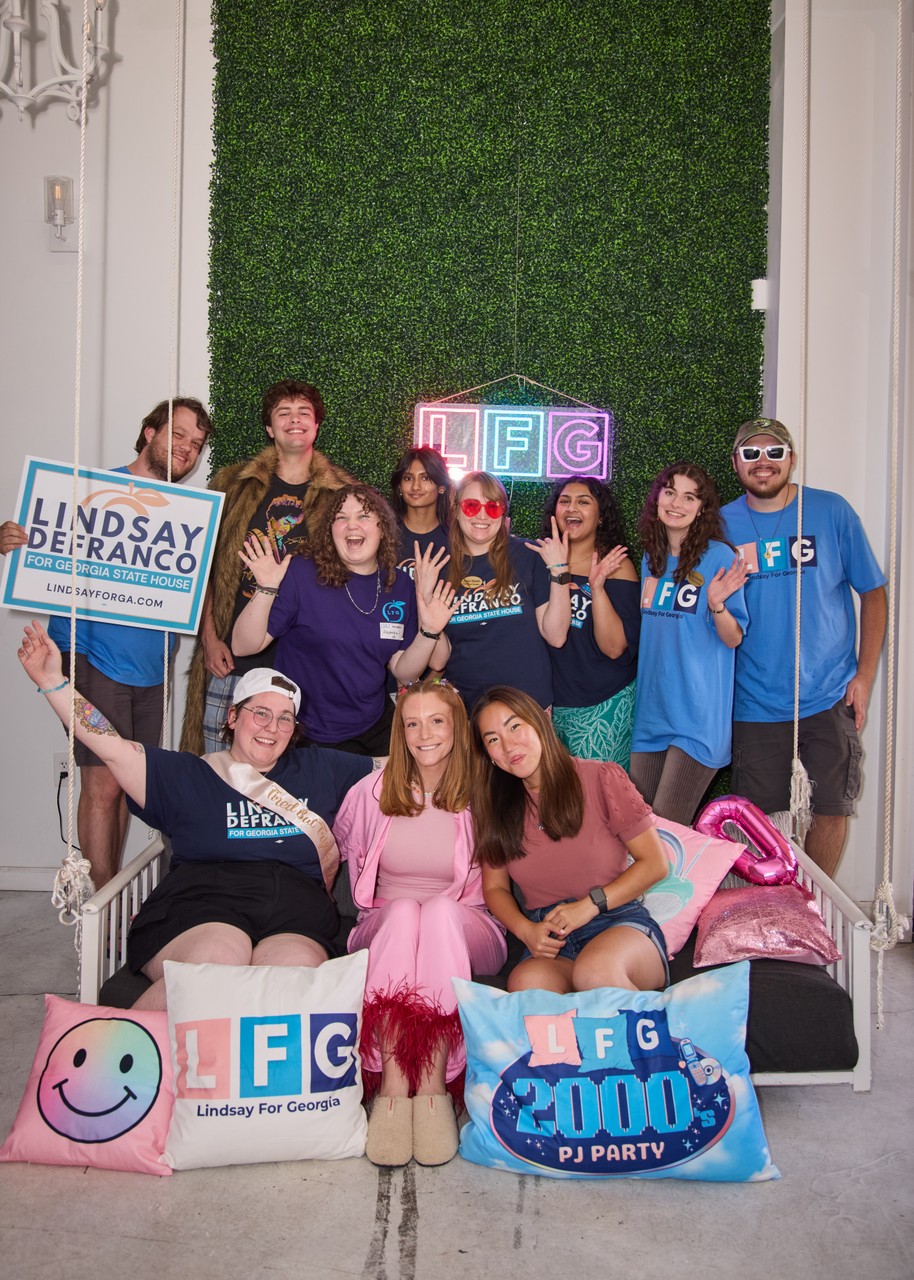 Group of people posing with signs and props, some wearing matching blue shirts, in front of a green wall.
