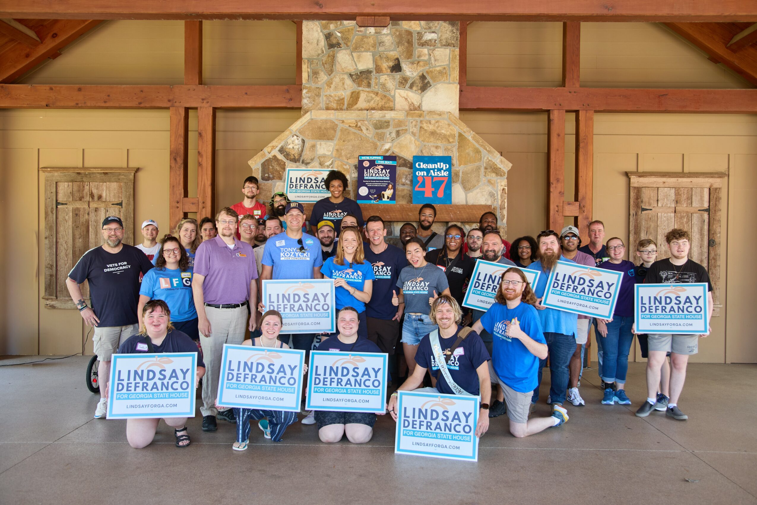 Group of people holding signs that read 'Lindsay Bercano' in a room with wooden beams and a stone fireplace.
