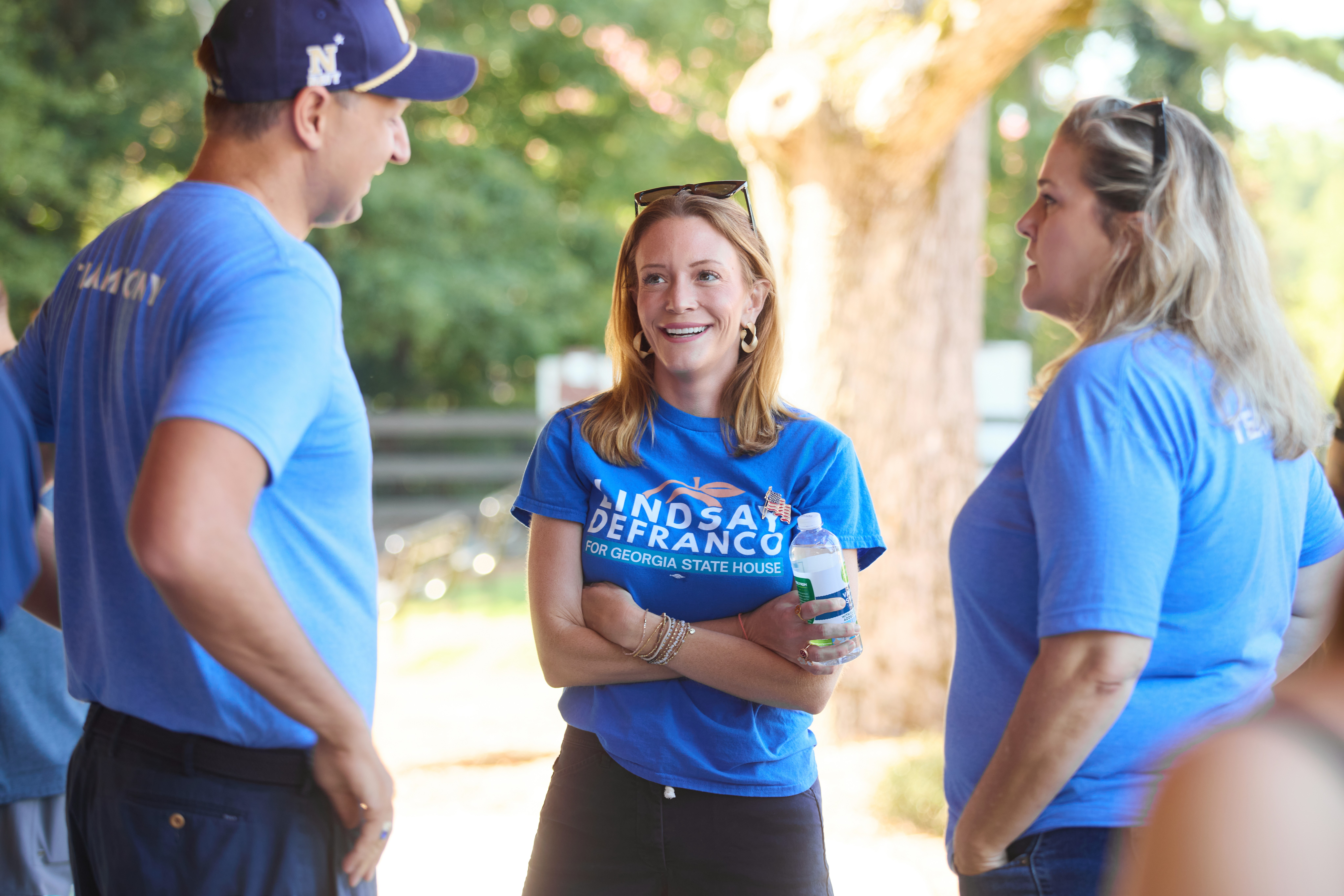 Three people in blue shirts talking outdoors, smiling, with trees and sunlight in the background.