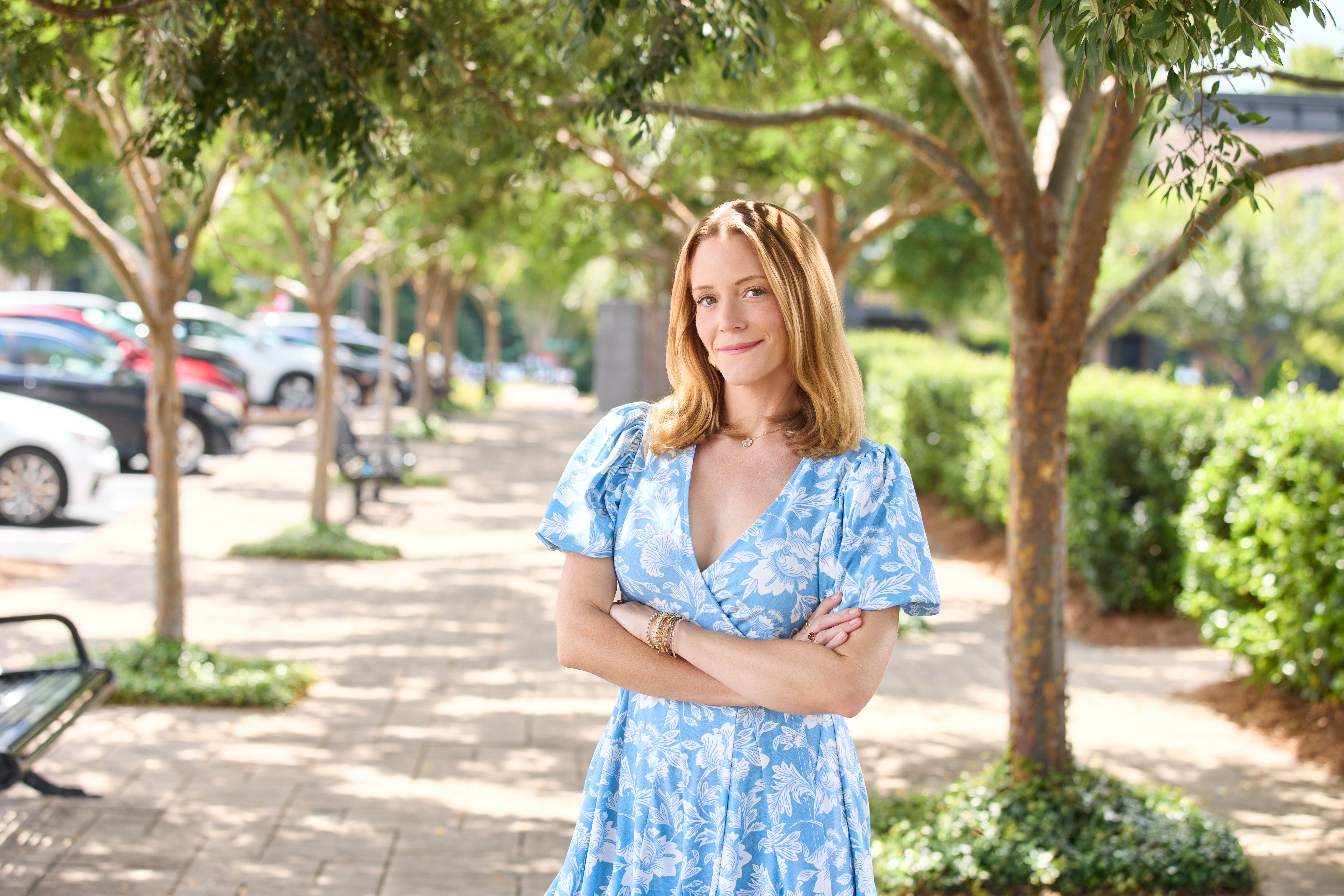 Woman with blonde hair in a blue floral dress standing outdoors on a sunny sidewalk, trees and parked cars in background.