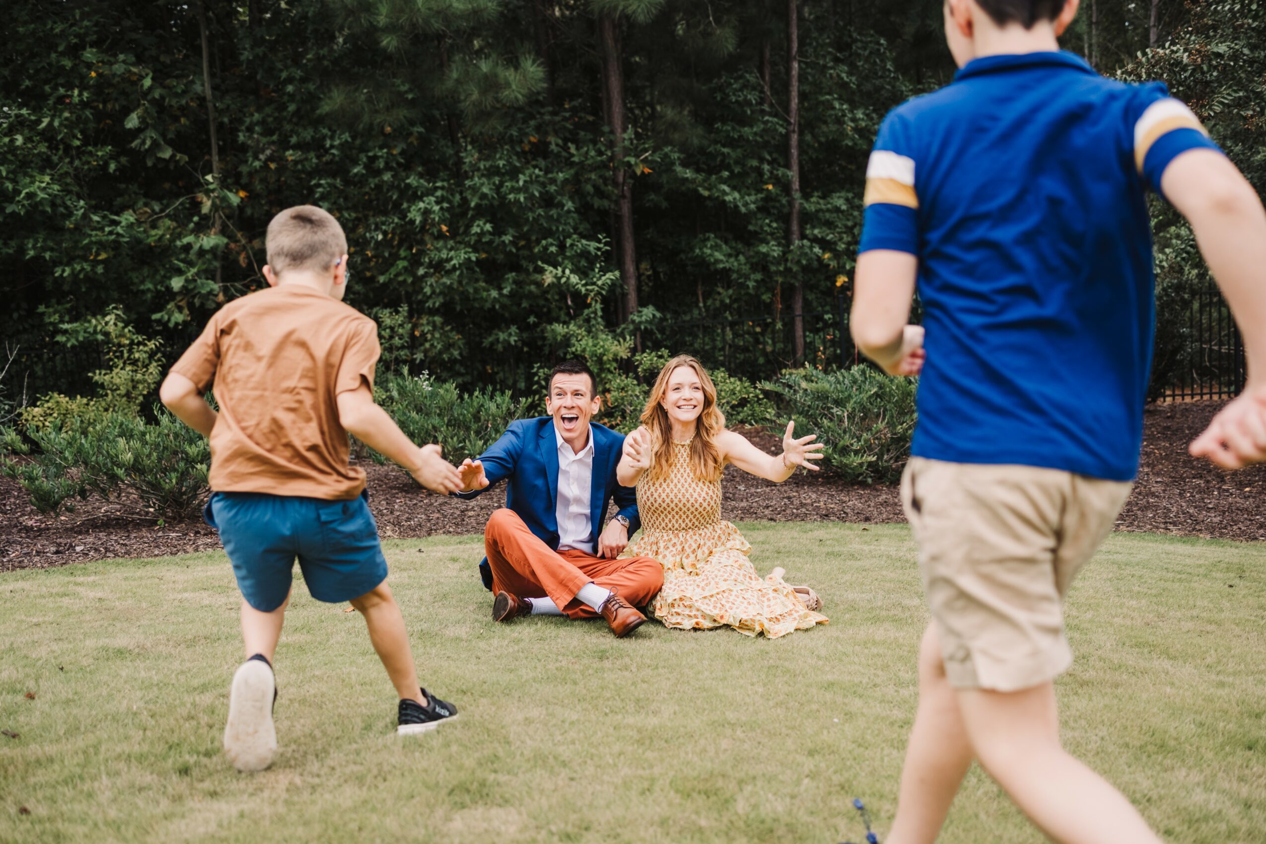 Two children run towards two people sitting on grass in a park, with trees in the background.