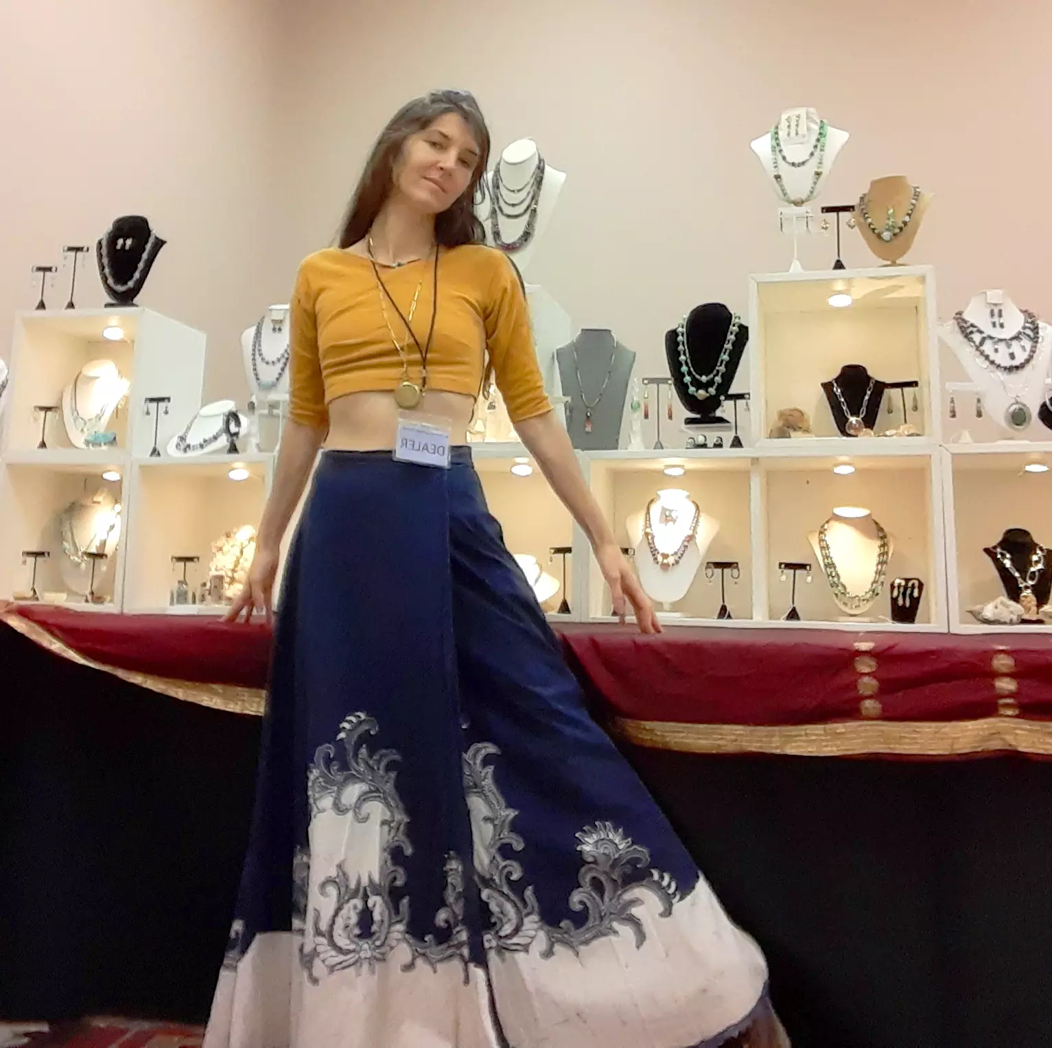 Young woman in yellow top and blue skirt with white pattern, standing in front of jewelry display, smiling, holding the skirt.