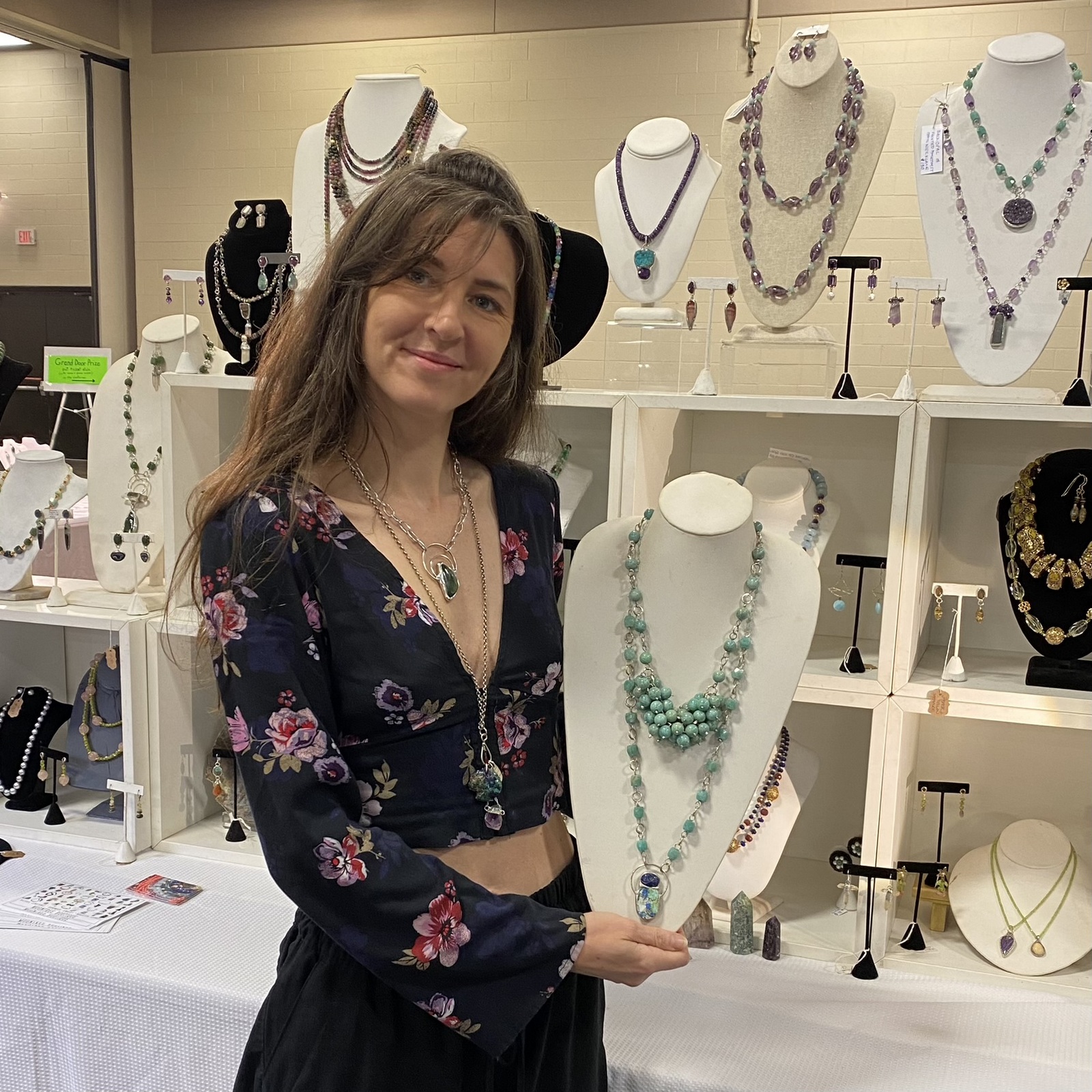 Young woman with long hair wearing a floral top, standing in jewelry store with necklaces displayed behind her.