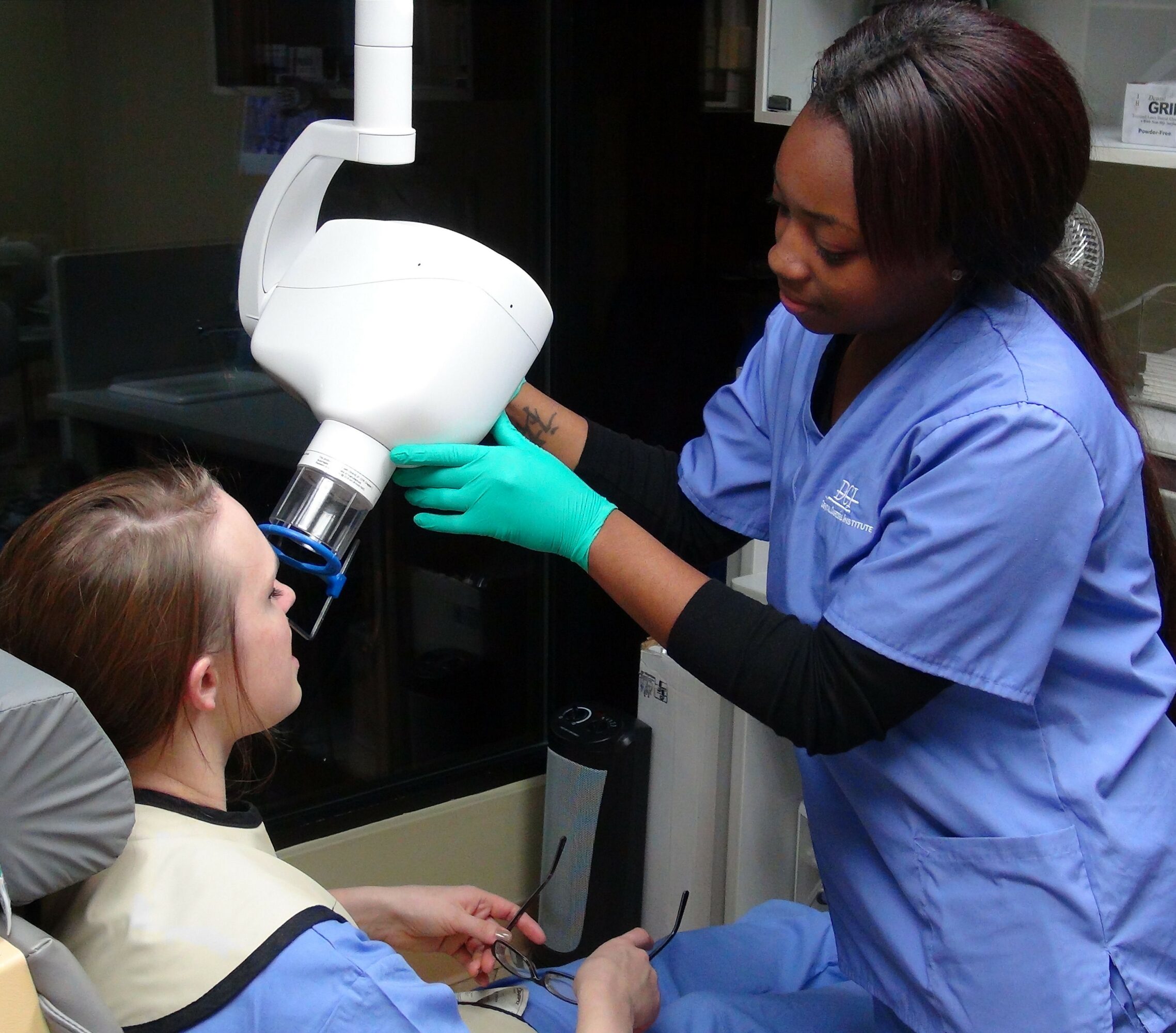 Dental professional in blue scrubs and gloves uses a microscope on a young patient in a dental chair.