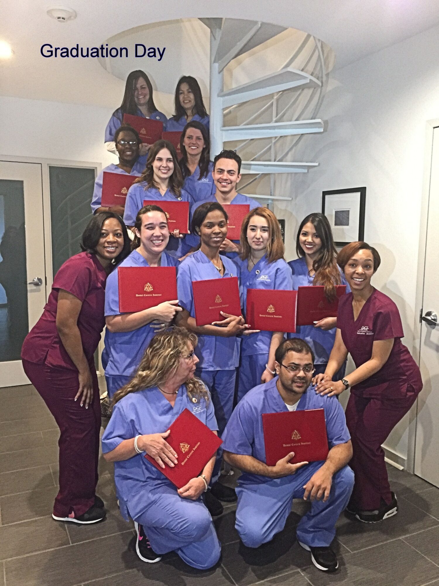 Group of graduates in scrubs holding diplomas, smiling, with a 'Graduation Day' sign in the background.