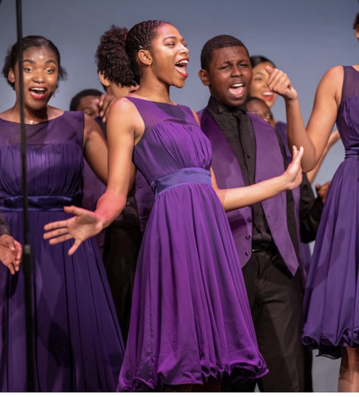 Group of children singing and dancing, wearing purple dresses and black suits, smiling and enjoying performance.