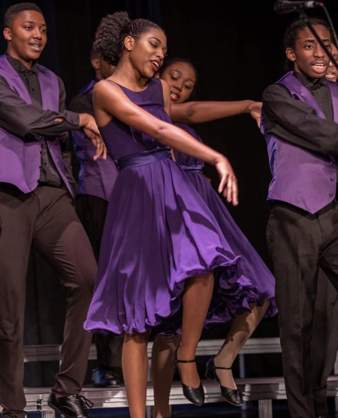 Group of young performers dancing on stage, wearing purple and black costumes, with joyful expressions.
