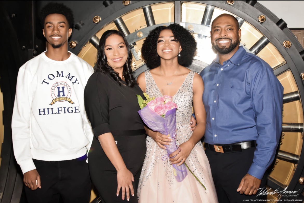 Four people standing together, smiling, with a large clock in the background, one holding a bouquet of flowers.