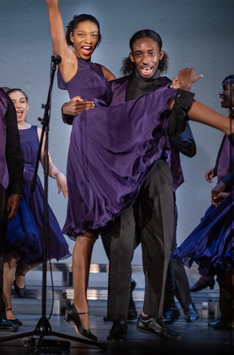 Group of people dancing on stage, wearing purple and black costumes, smiling and enjoying performance.