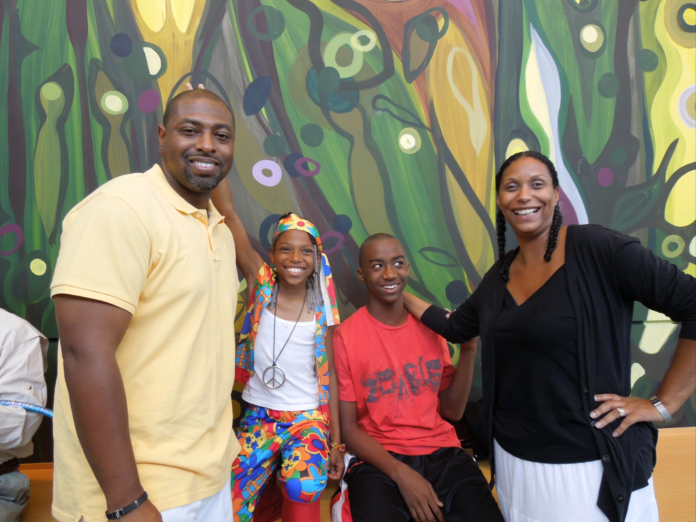 Four people smiling in front of a colorful abstract mural, two adults and two children, one girl and one boy.