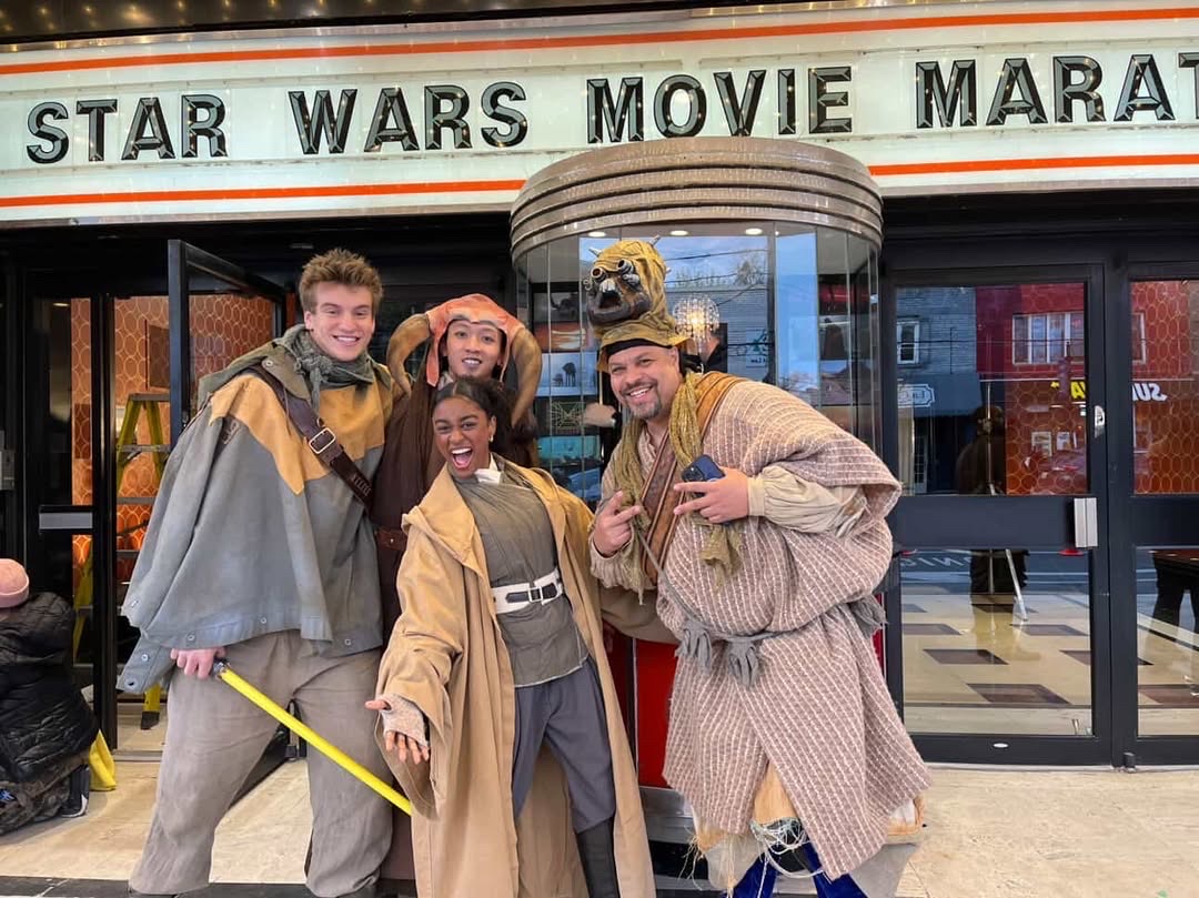 Four people dressed in costumes stand in front of a theater marquee that reads 'Star Wars Movie Marathon'.